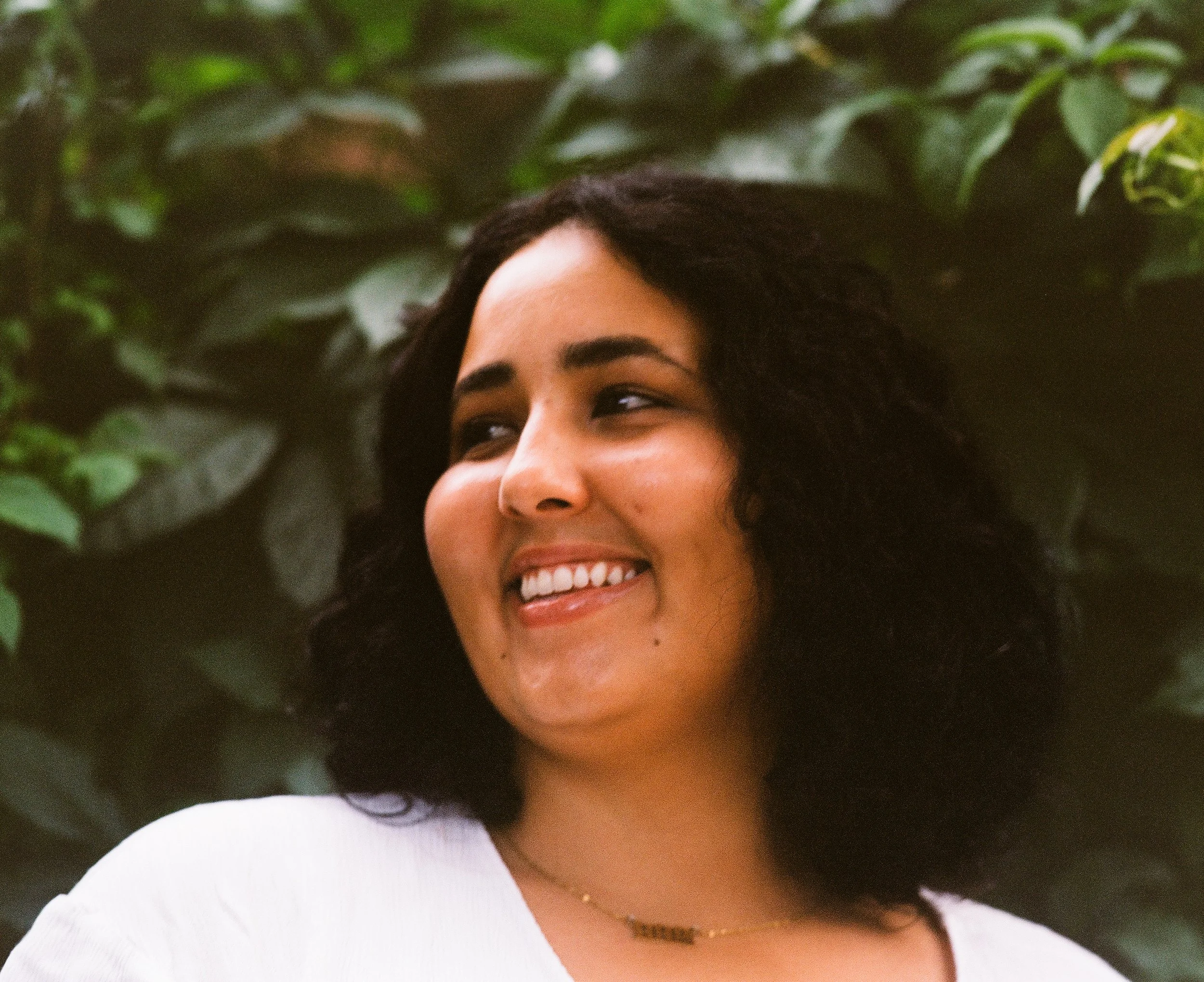 Close-up of a woman with dark curly hair smiling, wearing a white top and a gold necklace, with green foliage in the background.