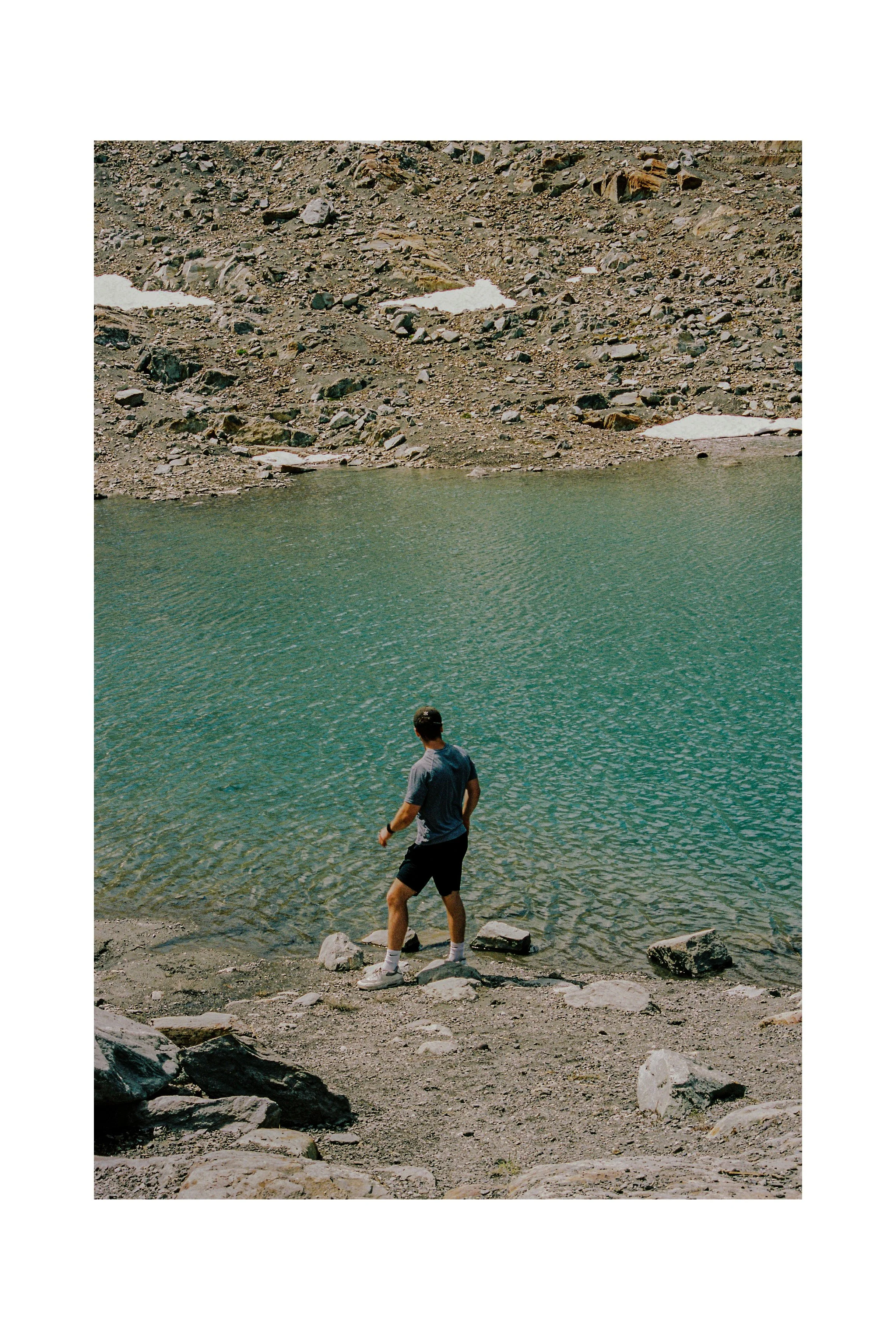 A person standing on a rocky lakeshore, looking at the water in a mountain setting with rocky terrain and patches of snow. Whistler, BC. 