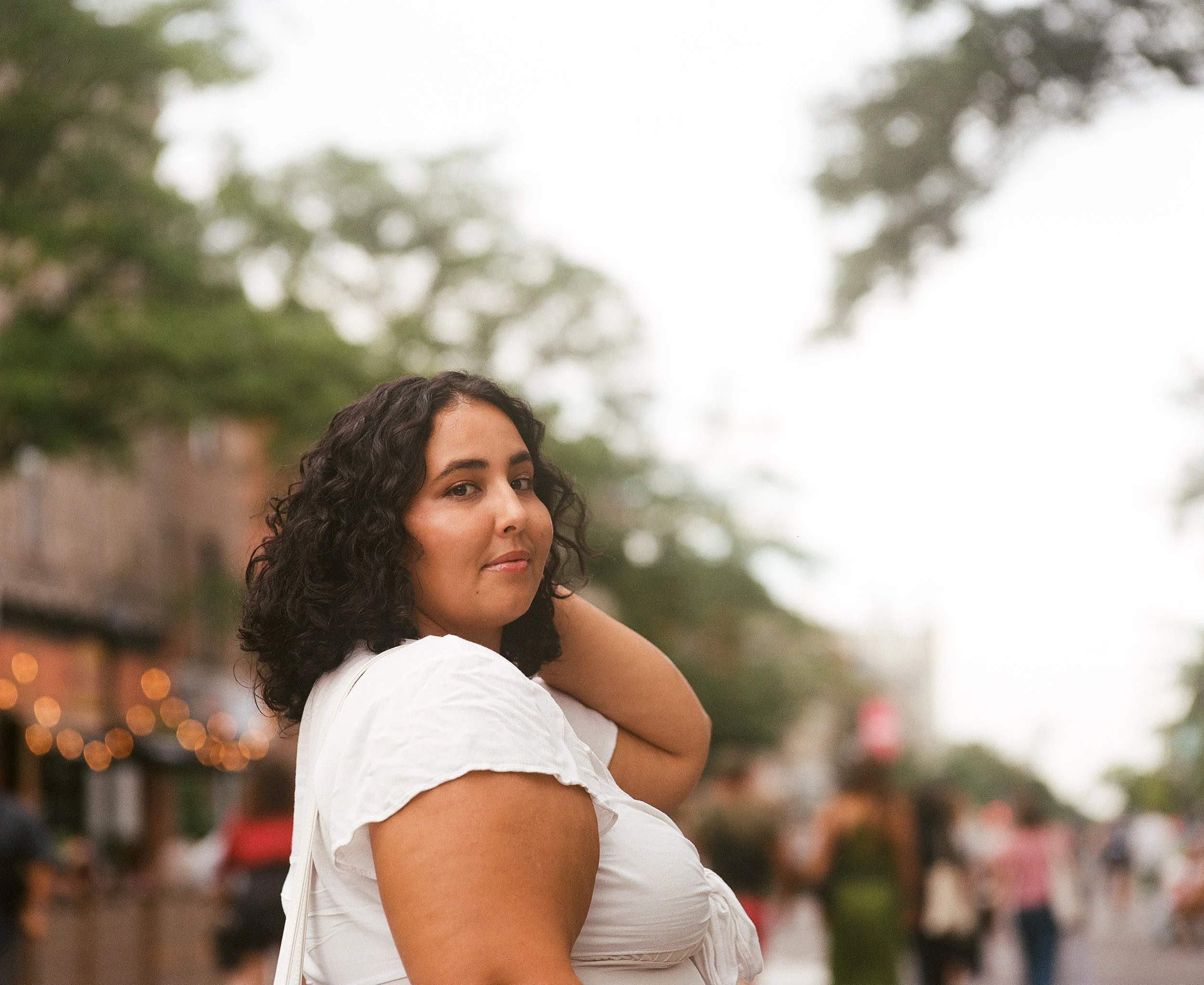 A woman with shoulder-length curly dark hair and medium skin tone in a white shirt stands outdoors, turning slightly to her left with her right hand resting on the back of her neck, on a lively street with trees and blurred people in the background.
