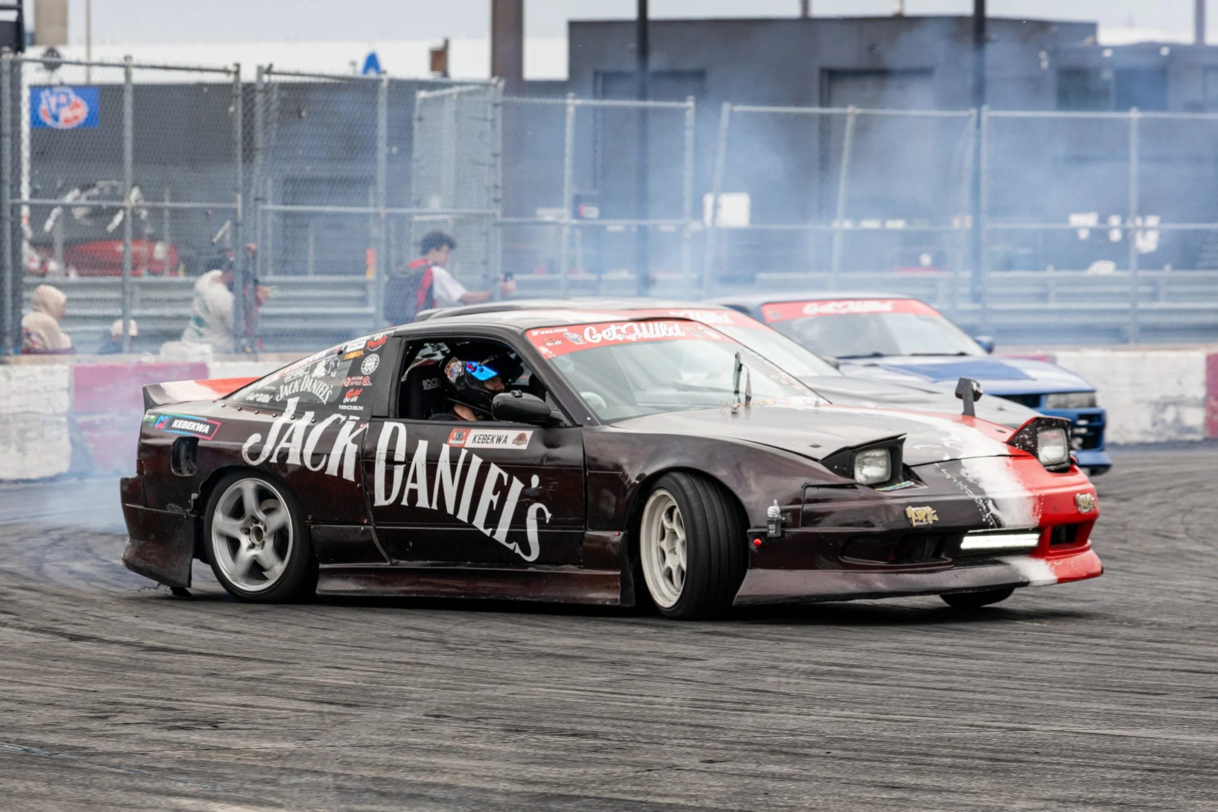 A black and red sports car with Jack Daniel's branding on the side performing a drift on the track, creating smoke from the tires. In the background, another car, a person walking, and a fence with people are visible.