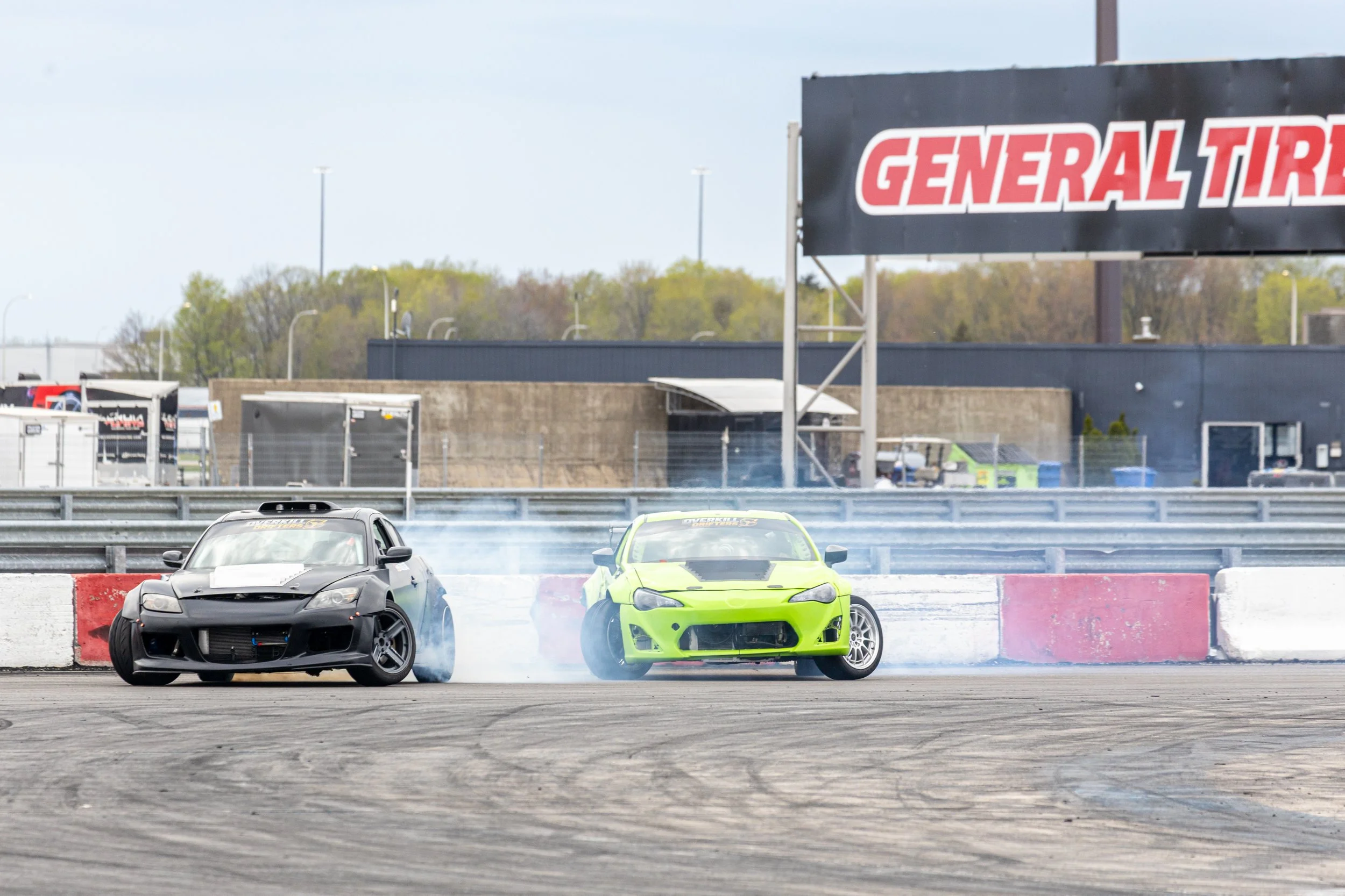 Two race cars, one black and one neon green, drifting on a track with smoke trailing behind. In the background, there is a sign that reads 'GENERALTIRE' and some trees.