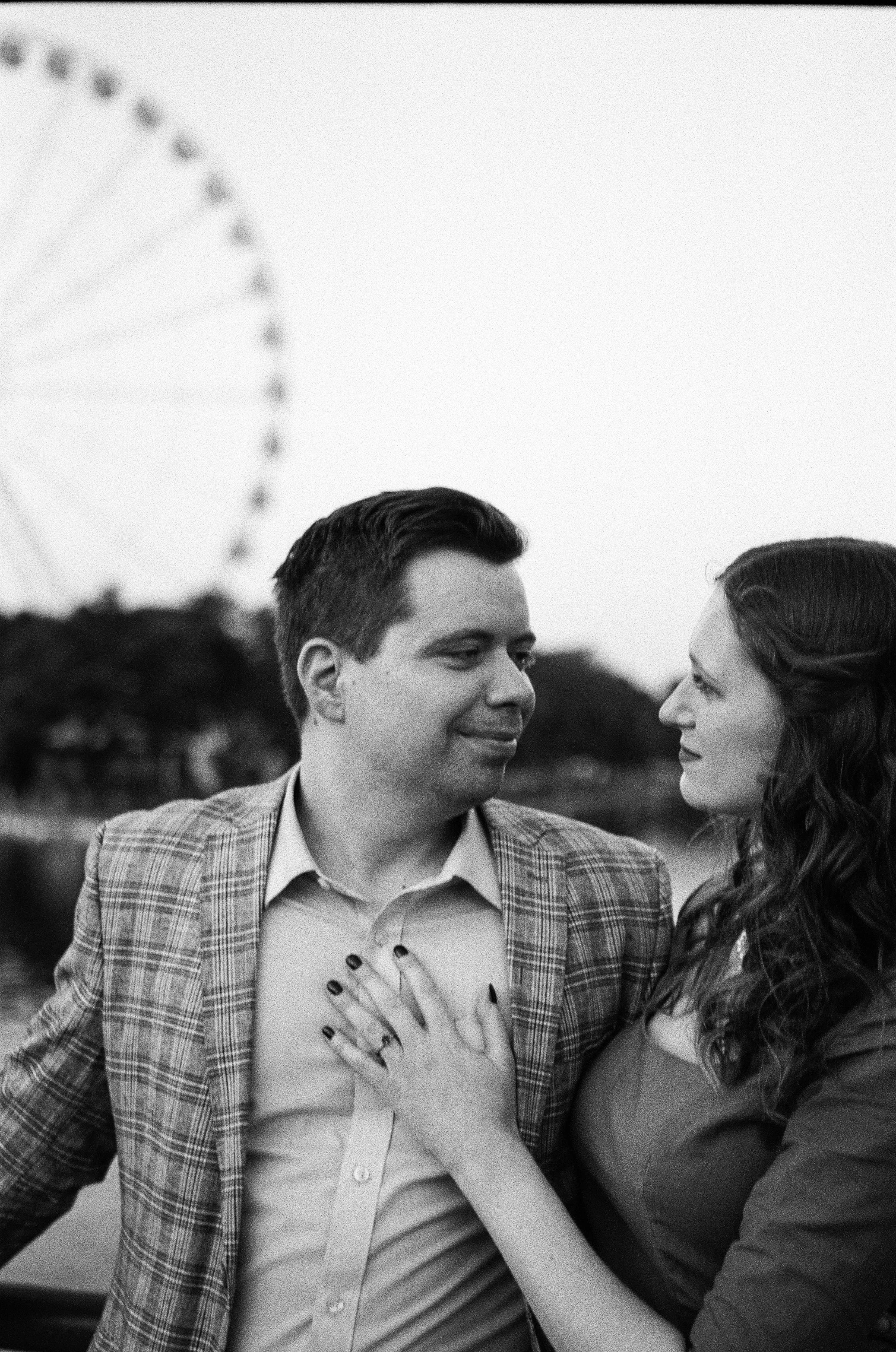 A black and white photo of a couple looking at each other, with a Ferris wheel in the background. Located in Montreal, Quebec. 