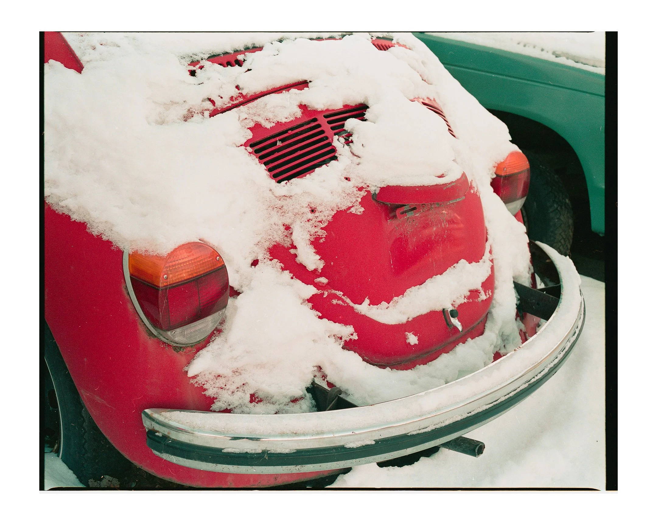 The back of a red vintage Volkswagen Beetle car covered with snow, with part of a green vehicle visible next to it.