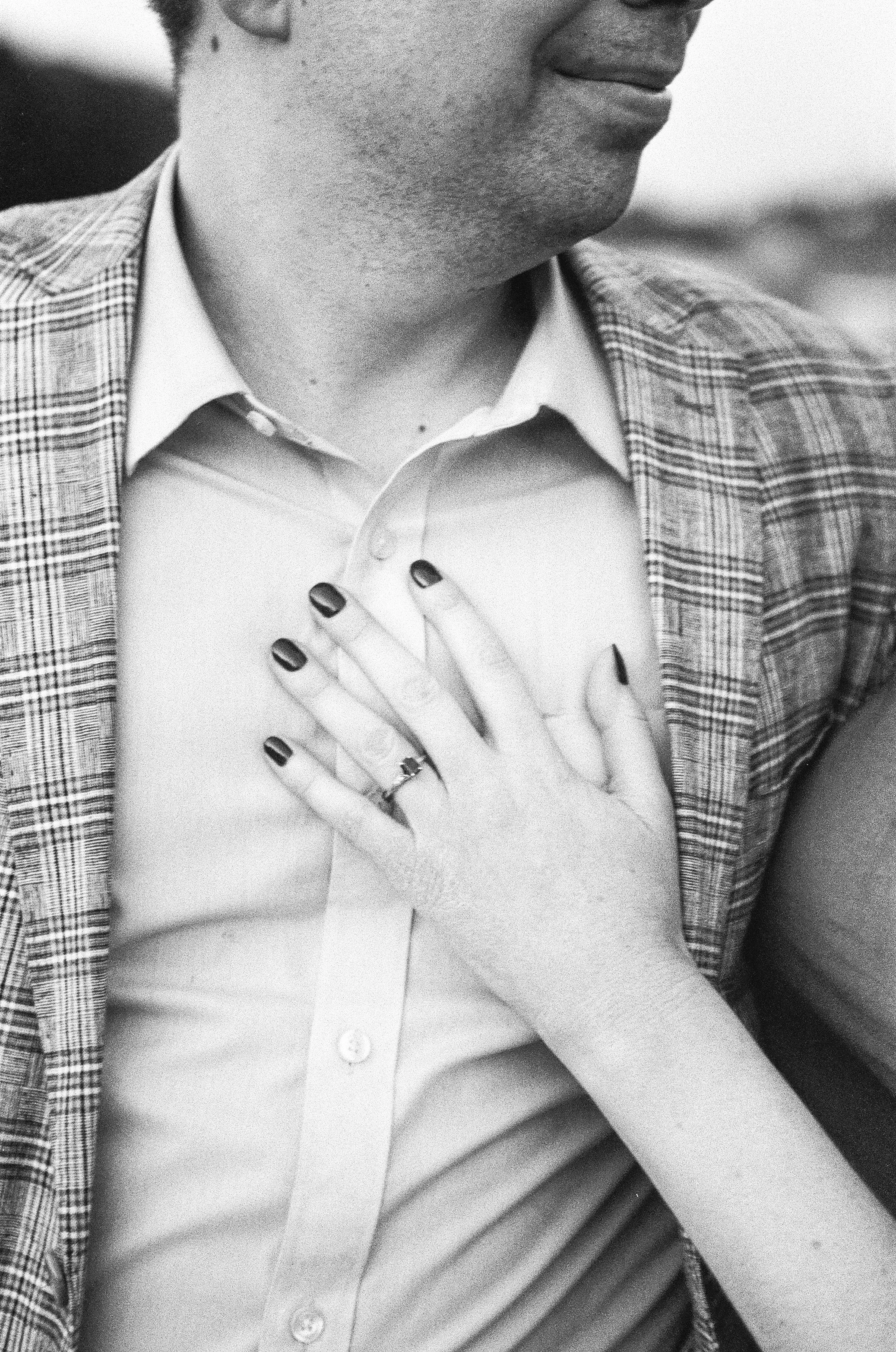 A woman with manicured nails and a wedding ring places her hand on a man's chest, who is dressed in a button-up shirt and a checkered blazer.