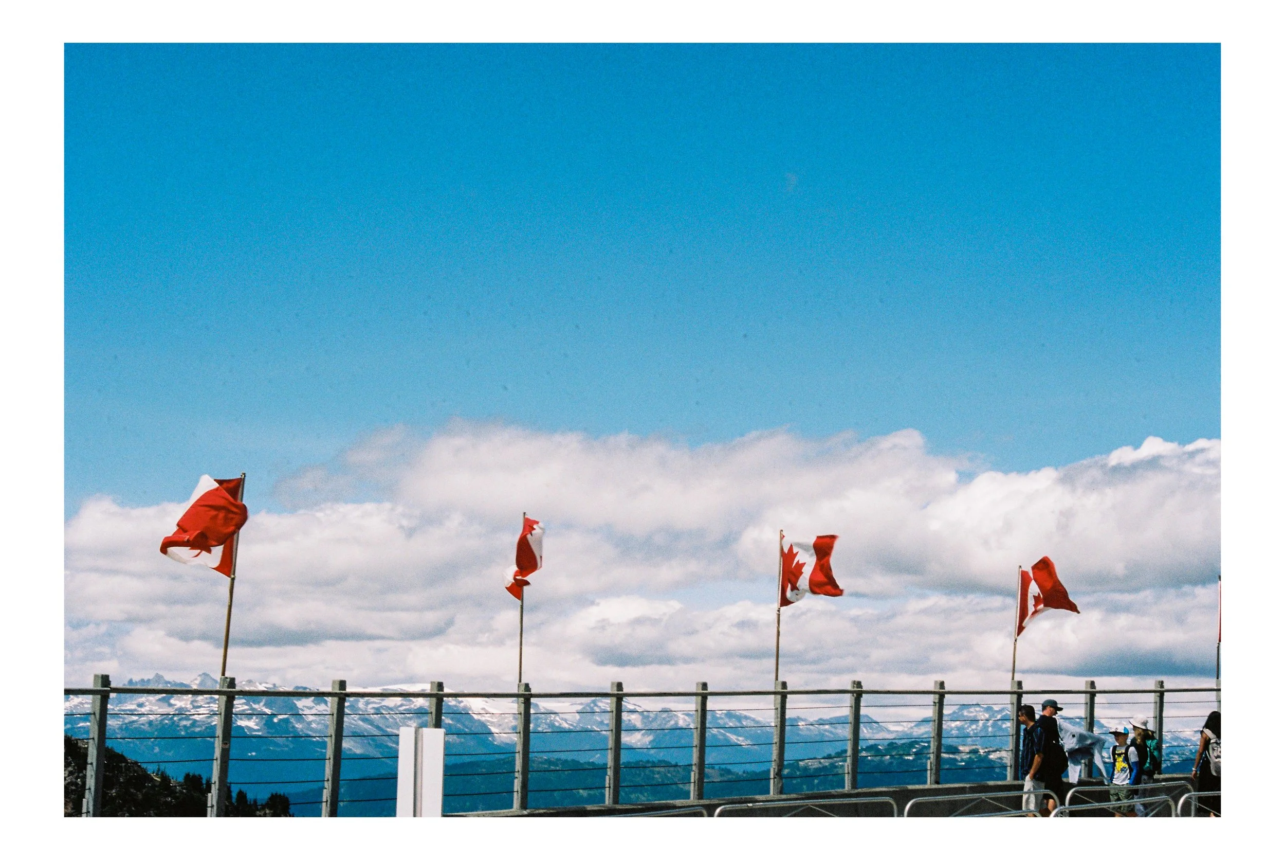 Several Canadian flags waving on flagpoles against a backdrop of snow-capped mountains and a partly cloudy sky, with a few people walking on a viewing platform in Whistler, BC.
