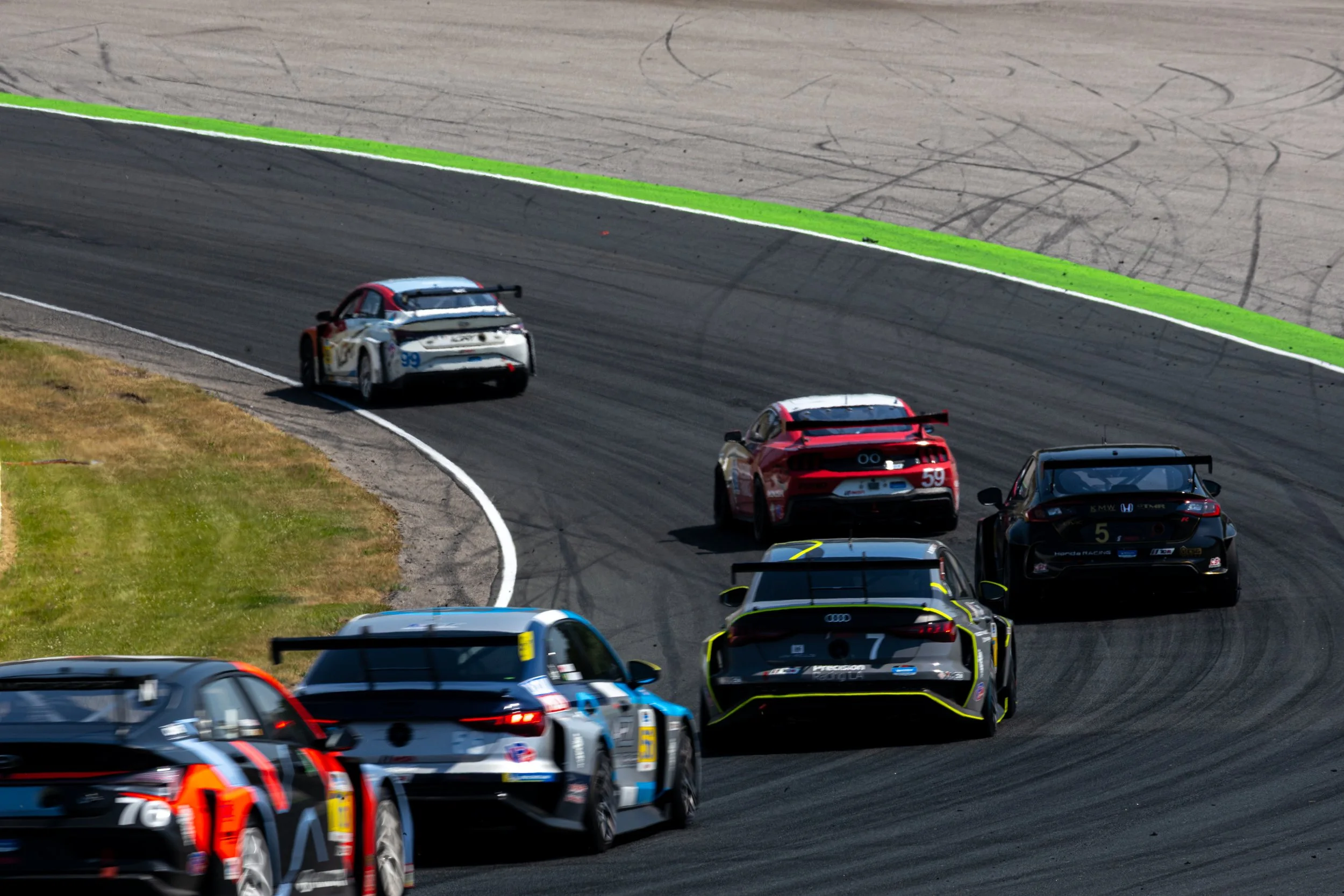 Multiple IMSA TCR class race cars at the Canadian Tire motorsports park, turning a corner on the asphalt with tire marks, with green painted curbs and grassy area beside the track.