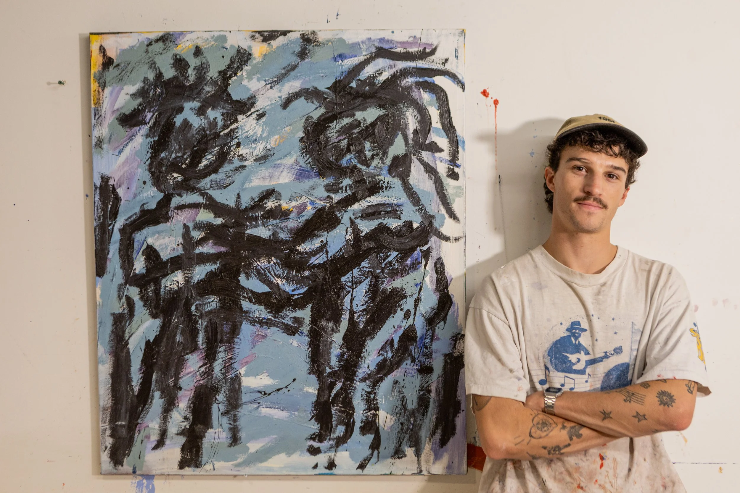 An artist stands with arms crossed next to an abstract painting of a horse on a white wall. The artist wears a beige cap, a white T-shirt with a blue guitar and musical notes graphic, and has tattoos on his arms.