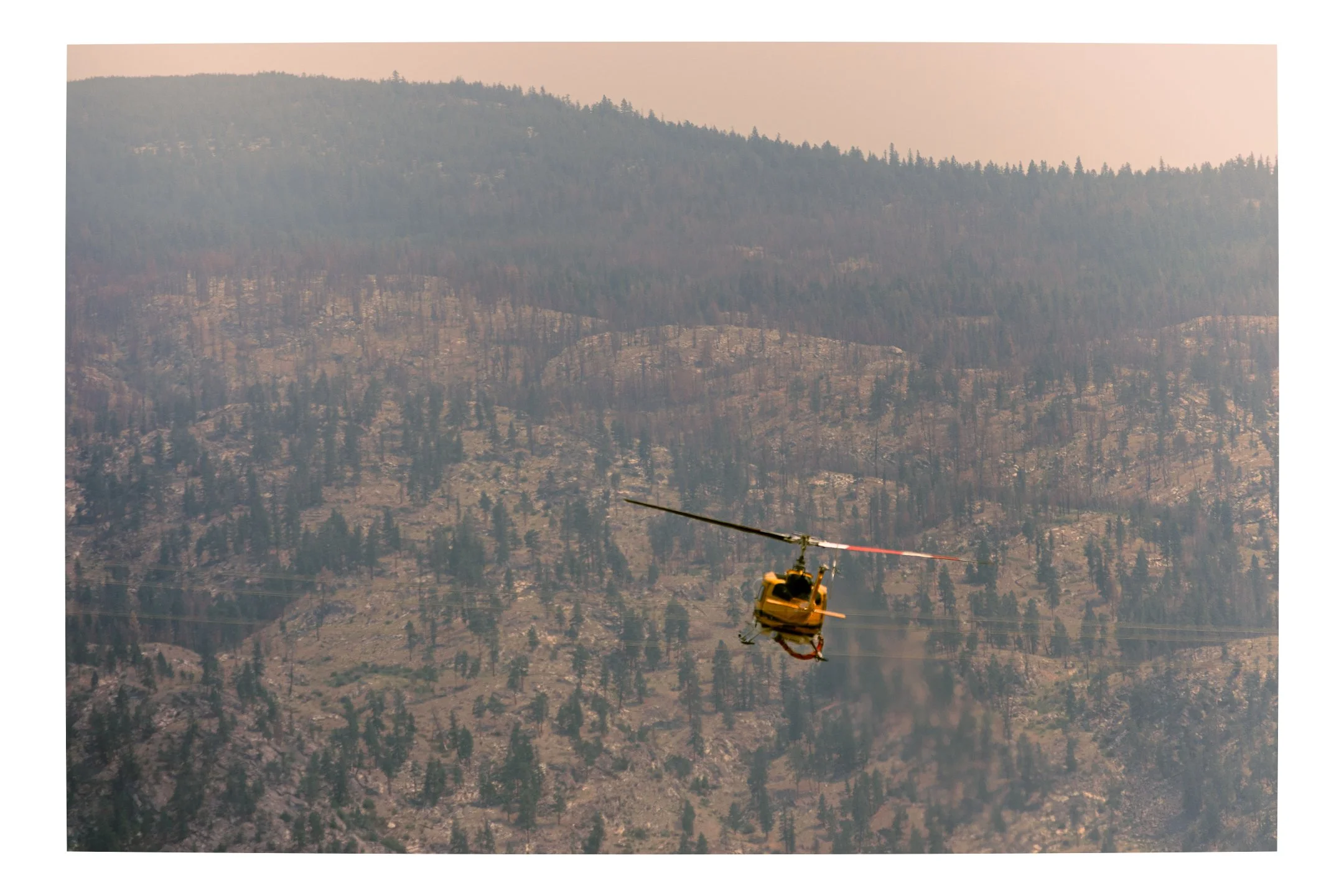 Yellow  Canadian forest fire helicopter flying over forested mountains with smoke in the background.