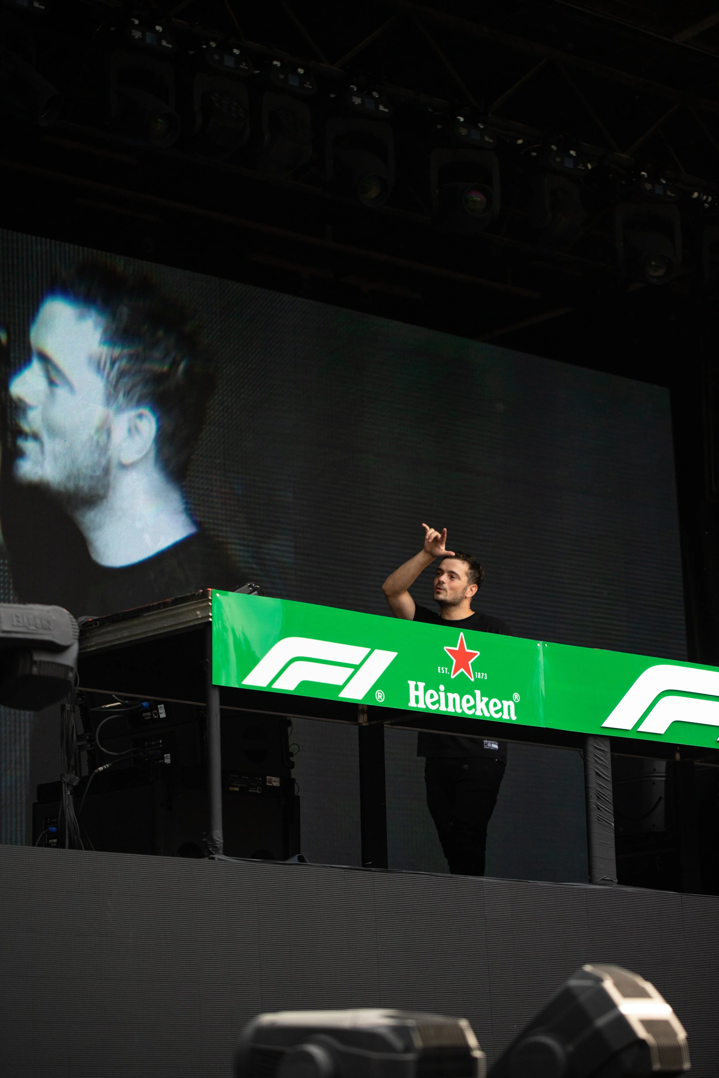 Martin Garrix is performing at a music event, standing behind a green Heineken-sponsored DJ booth with the Formula 1 logo on it. There is a large screen behind him showing a close-up two shot of his face.