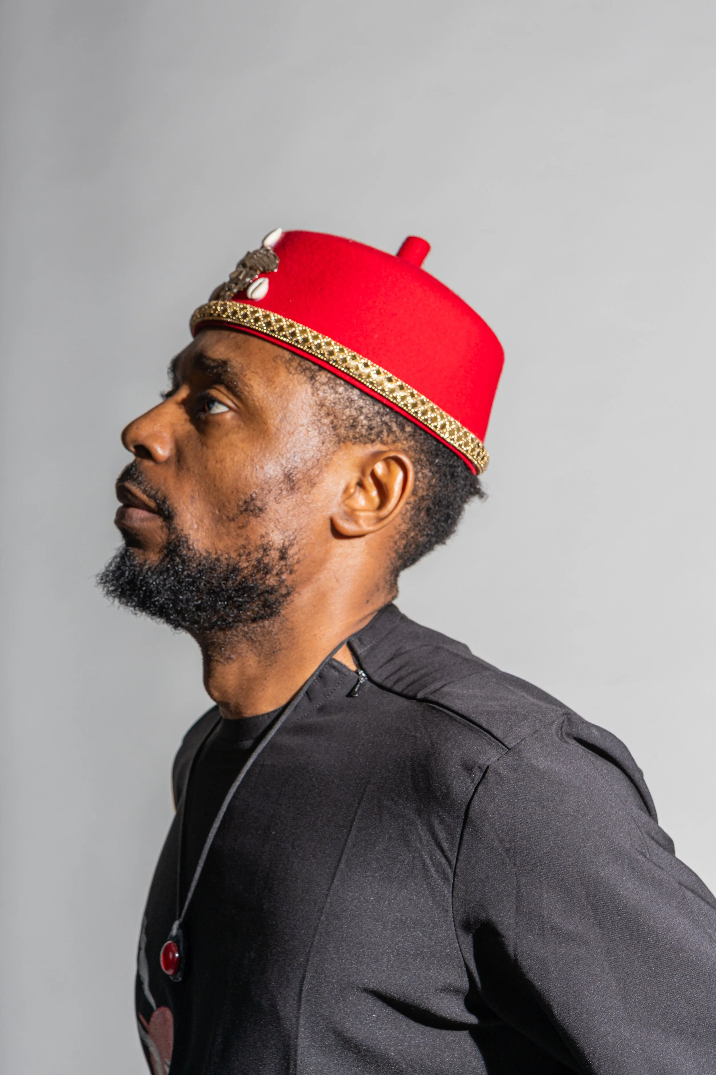 Man in profile wearing a red fez hat with gold trim and decorative embellishments, against a plain background.