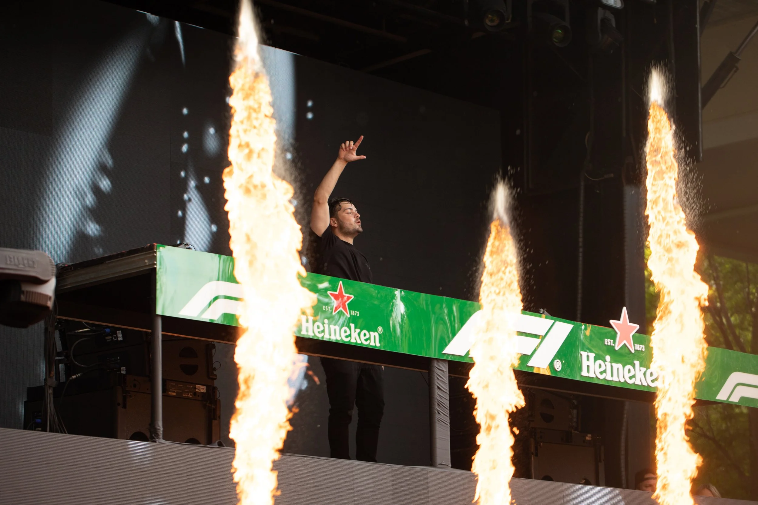Martin Garrix standing behind a Heineken-branded bar at a Formula 1 event, with flames shooting up in front of them, during a nighttime setting.