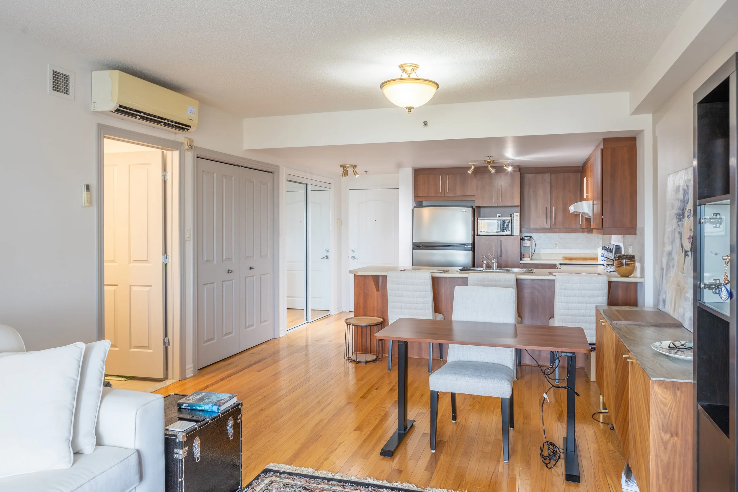 Living room with white sofa, wooden floor, and kitchen in background with brown cabinets and stainless steel appliances.