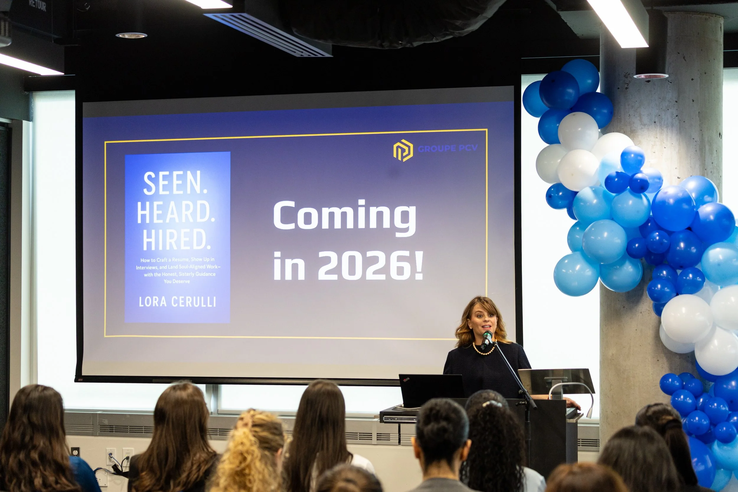 Lora giving a presentation at a Concordia JMSB conference with a large screen displaying the announcement 'Coming in 2026!' for her new novel. 