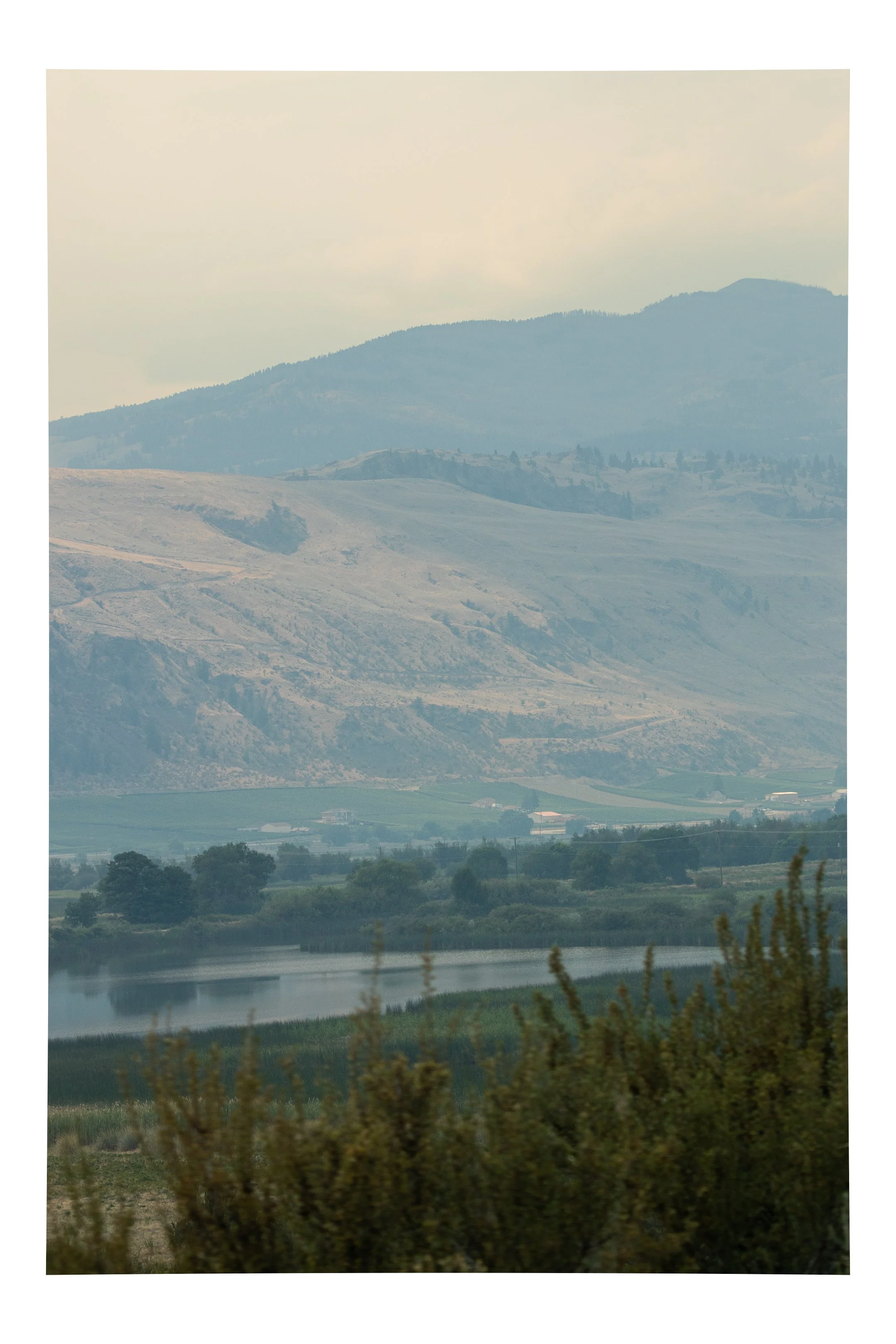 Scenic landscape featuring a river in the foreground, green shrubs, rolling hills, and mountains in the background under a smoky sky, caused by fires fires, 2022. Taken in Oliver, BC. 