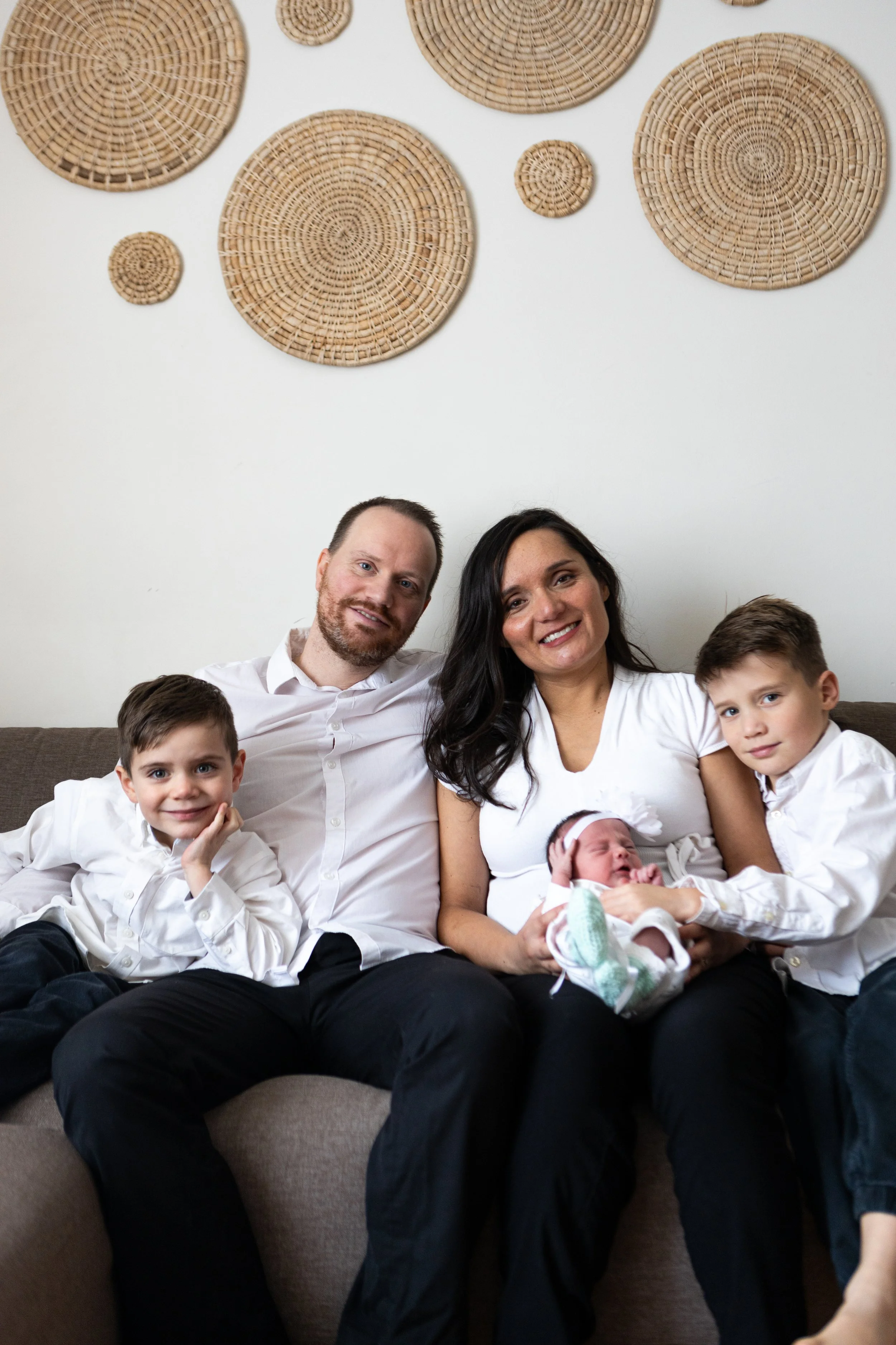 A family of five sitting on a couch, with two young boys, a woman holding a newborn, and a man smiling, with woven wall decorations behind them.