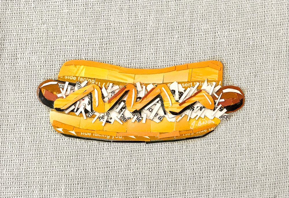 A hot dog-shaped patch made from small pieces of paper, with mustard, ketchup, and relish details, placed on a gray fabric background.