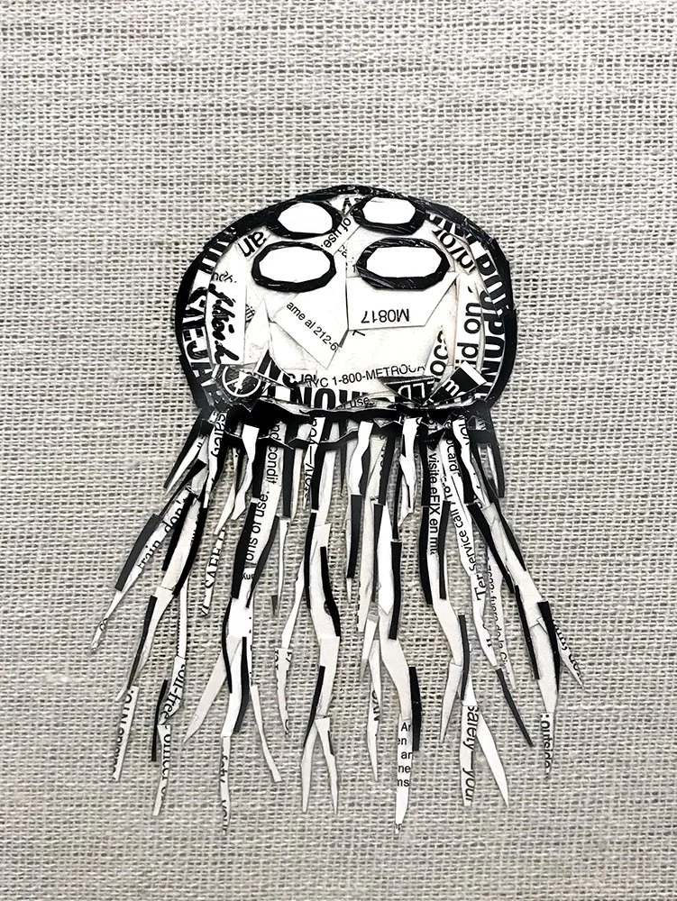 A paper collage of a jellyfish made from cut-out pieces of printed paper with tentacles hanging below, placed on a textile background.