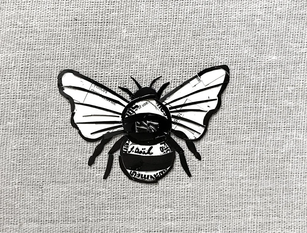 Decorative bee made from black cut-out paper and printed paper, mounted on a textured beige background.