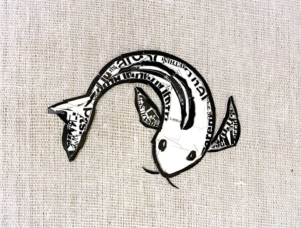 Paper cut-out of a cartoon fish, made from black and white newspaper or magazine strips, placed on a textured beige background.