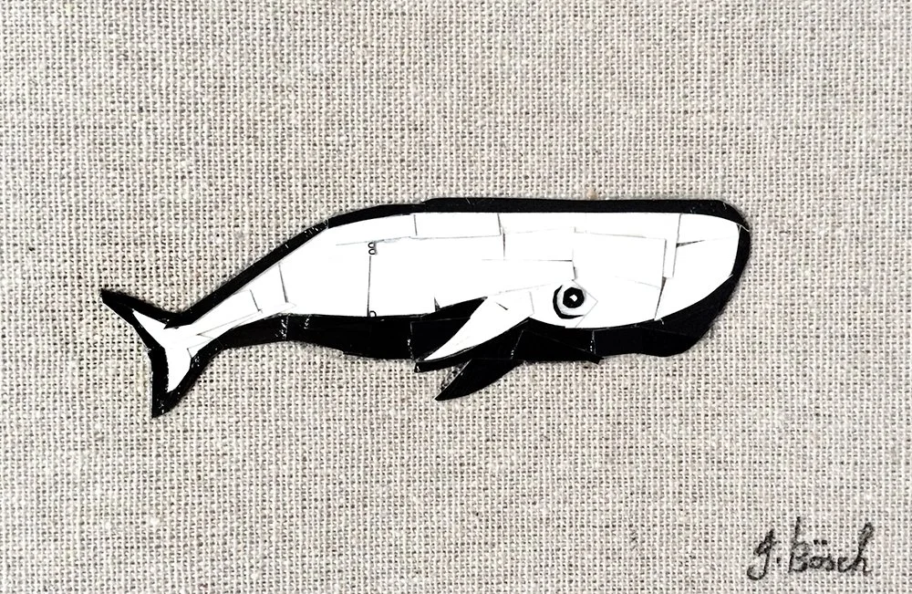 A black and white paper craft of a whale on a textured fabric background with the artist's signature at the bottom right.