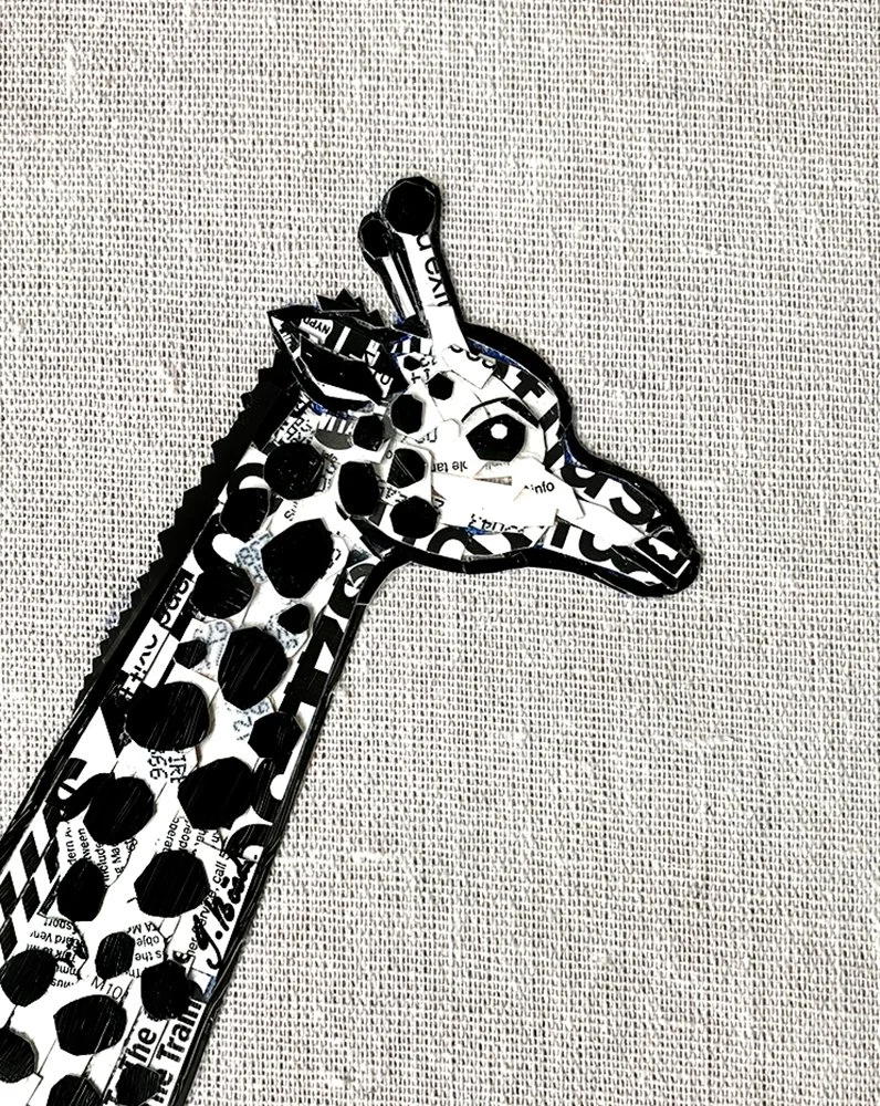 A metal sculpture of a giraffe made from cutouts of black and white printed paper, set against a textured light-colored fabric background.
