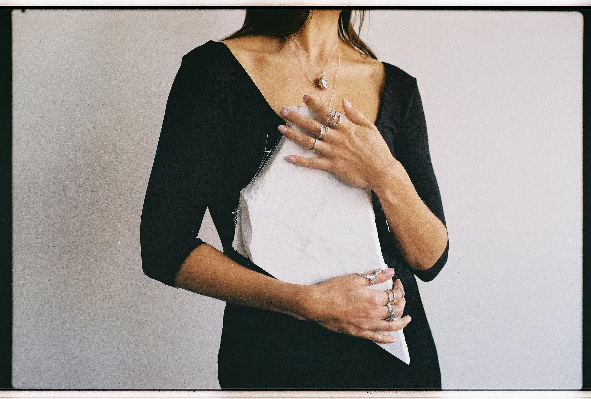 A woman in a black dress holding a white object against her chest with jewelry on her fingers, hands, and neck.