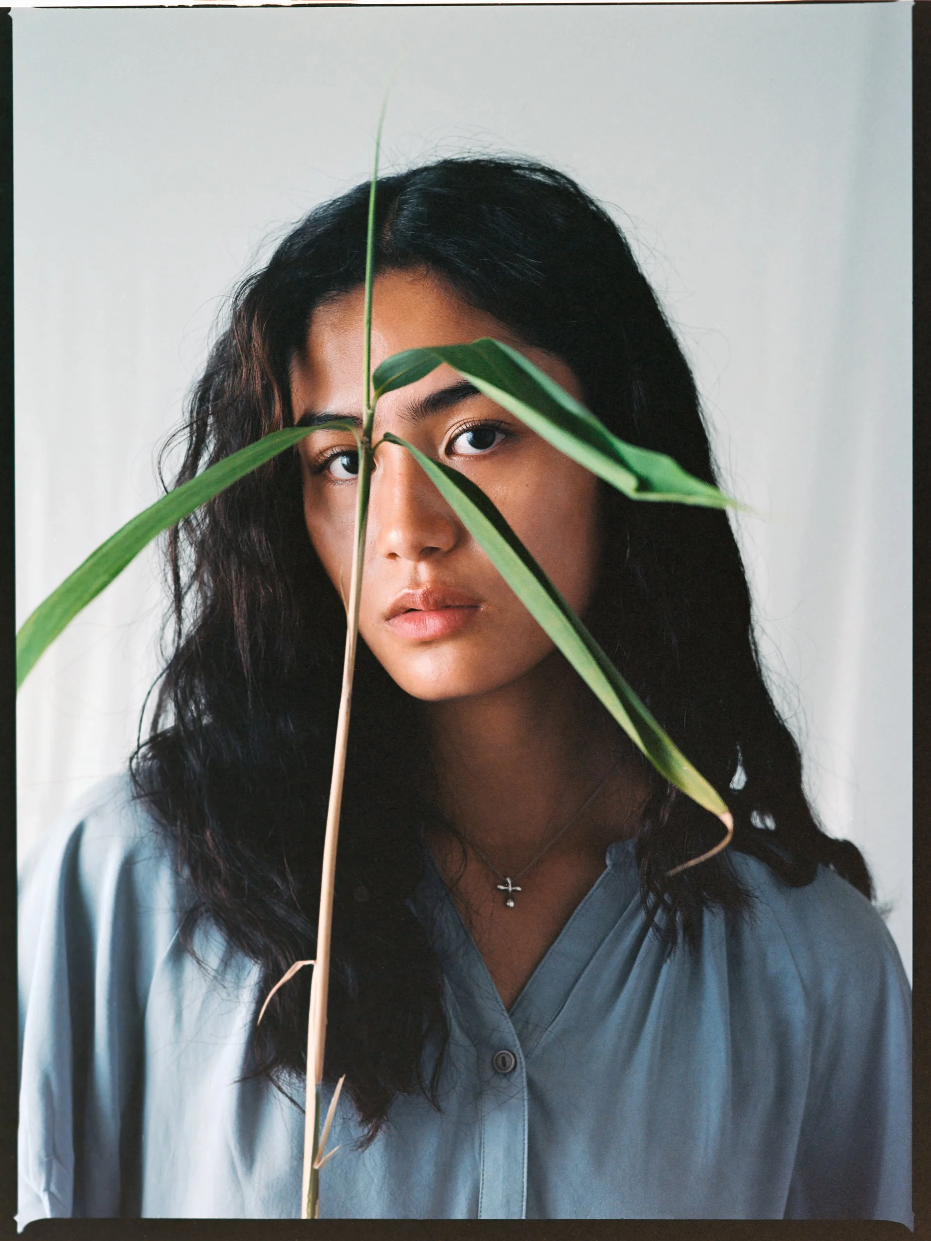 Young woman with dark hair and tan skin, wearing a light blue shirt, partially obscured face by a large green leaf.