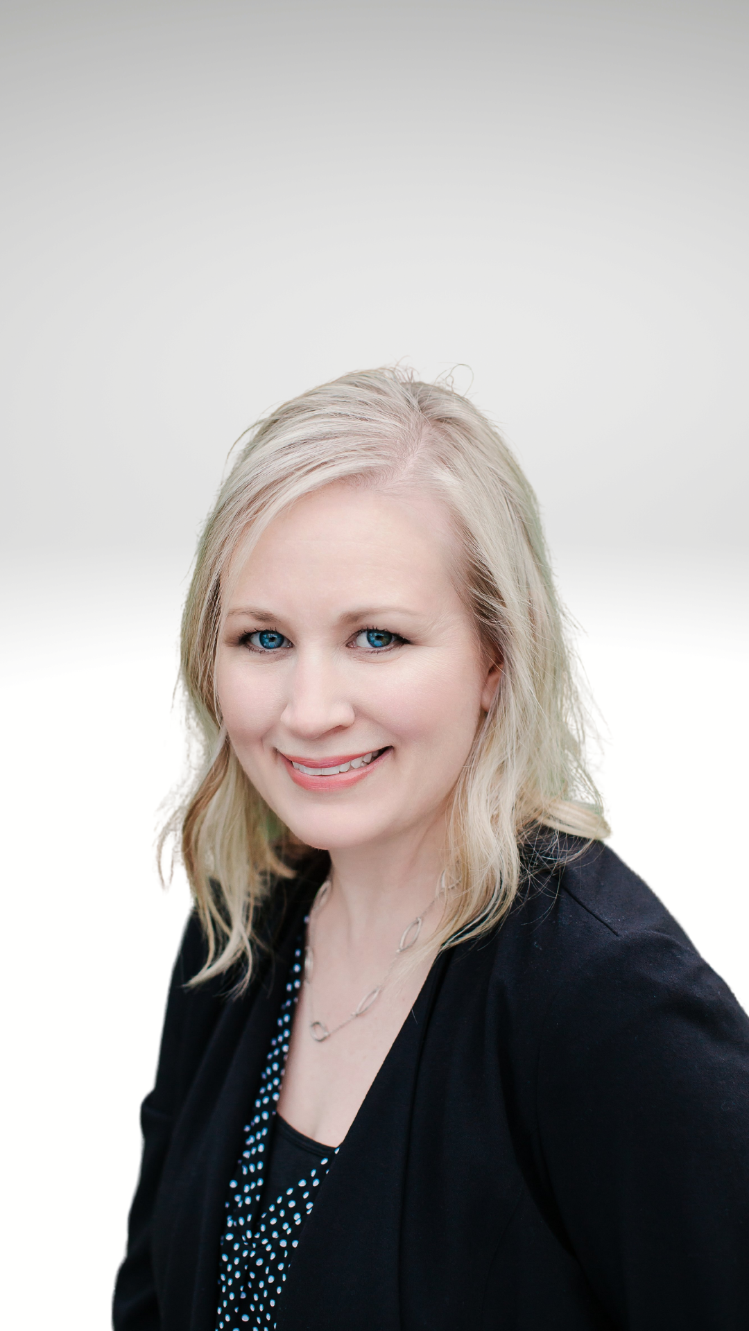 Portrait of a woman with blonde hair and blue eyes, smiling, wearing a black blazer and a black and white polka dot top, against a white background.