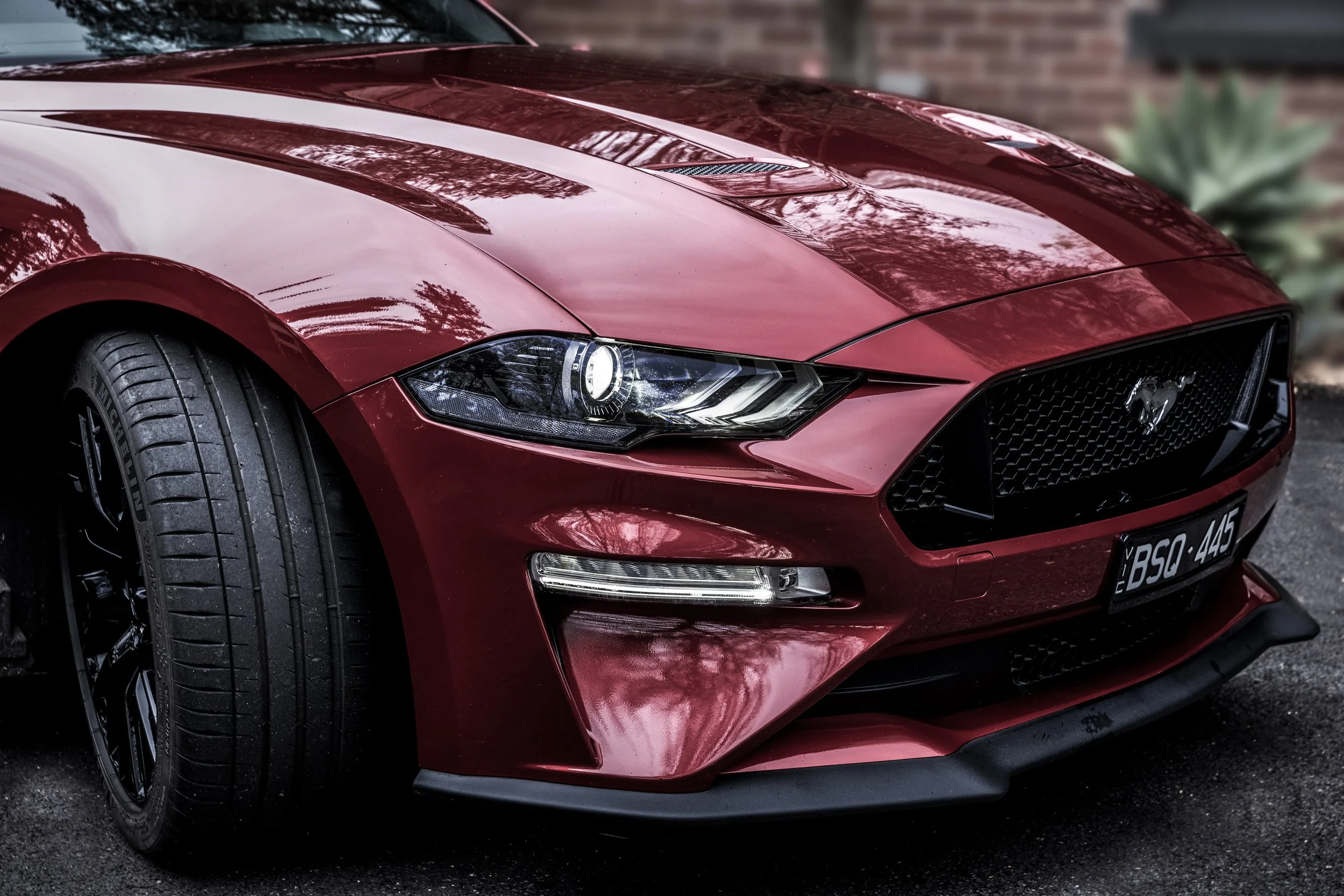 Close-up of a red Ford Mustang sports car showing the front left side, including the headlight, grille with Mustang emblem, and part of the front bumper and tire.