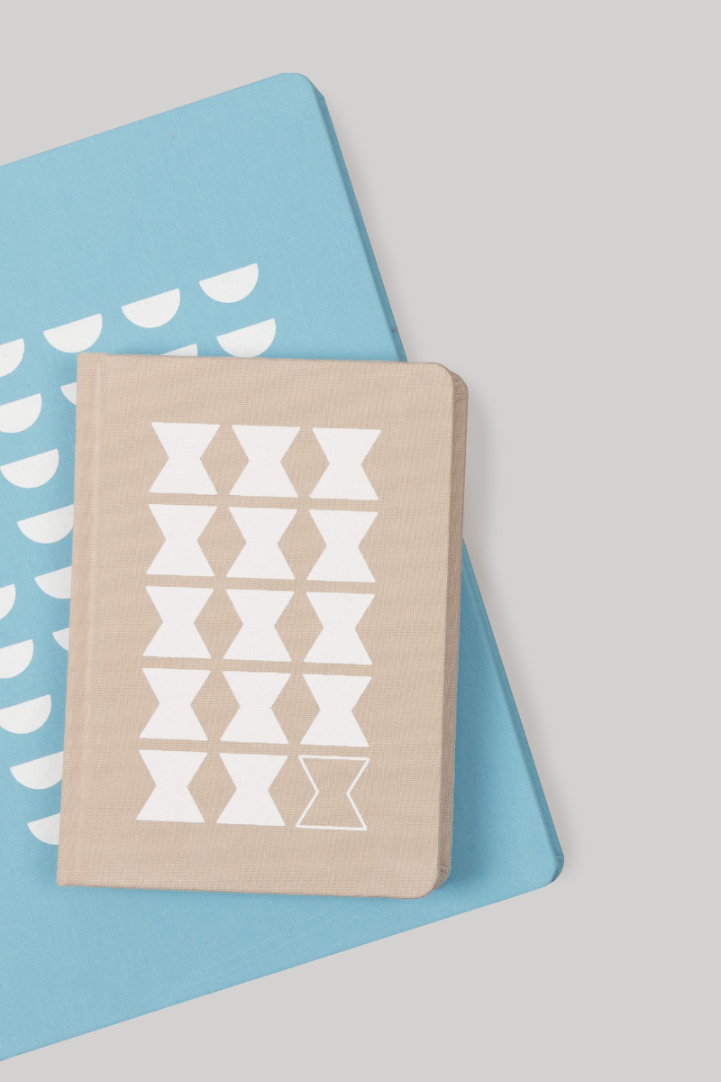 Two notebooks, one beige with white geometric hourglass patterns and one blue with white semi-circle patterns, laid on top of each other on a light gray background.