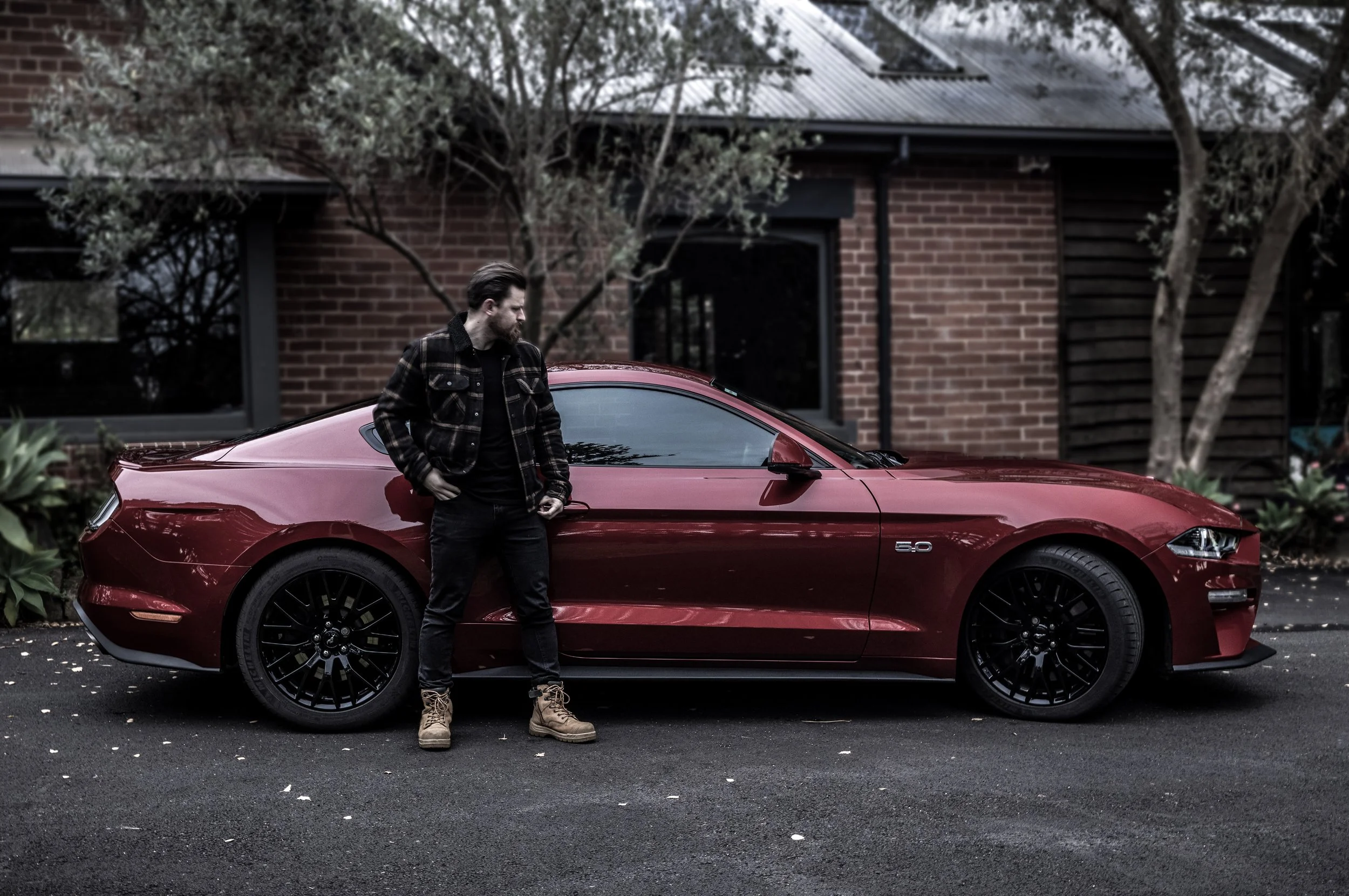 A man in a black jacket and tan boots standing next to a red sports car in front of a house with brick walls and trees.