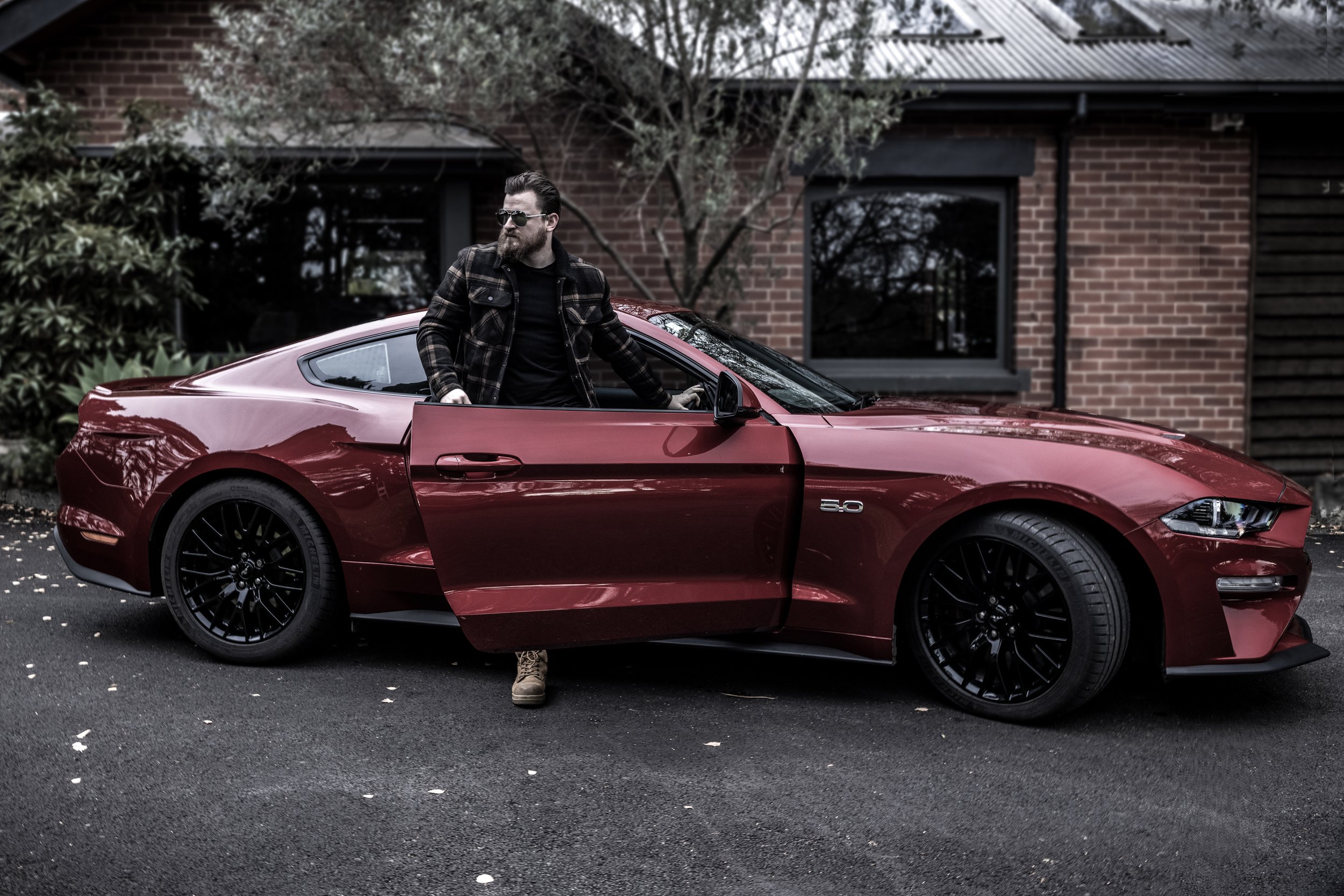 A man with sunglasses and a beard, wearing a plaid jacket and beige shoes, stands by a red sports car with one door open in front of a house with brick walls and dark-framed windows.