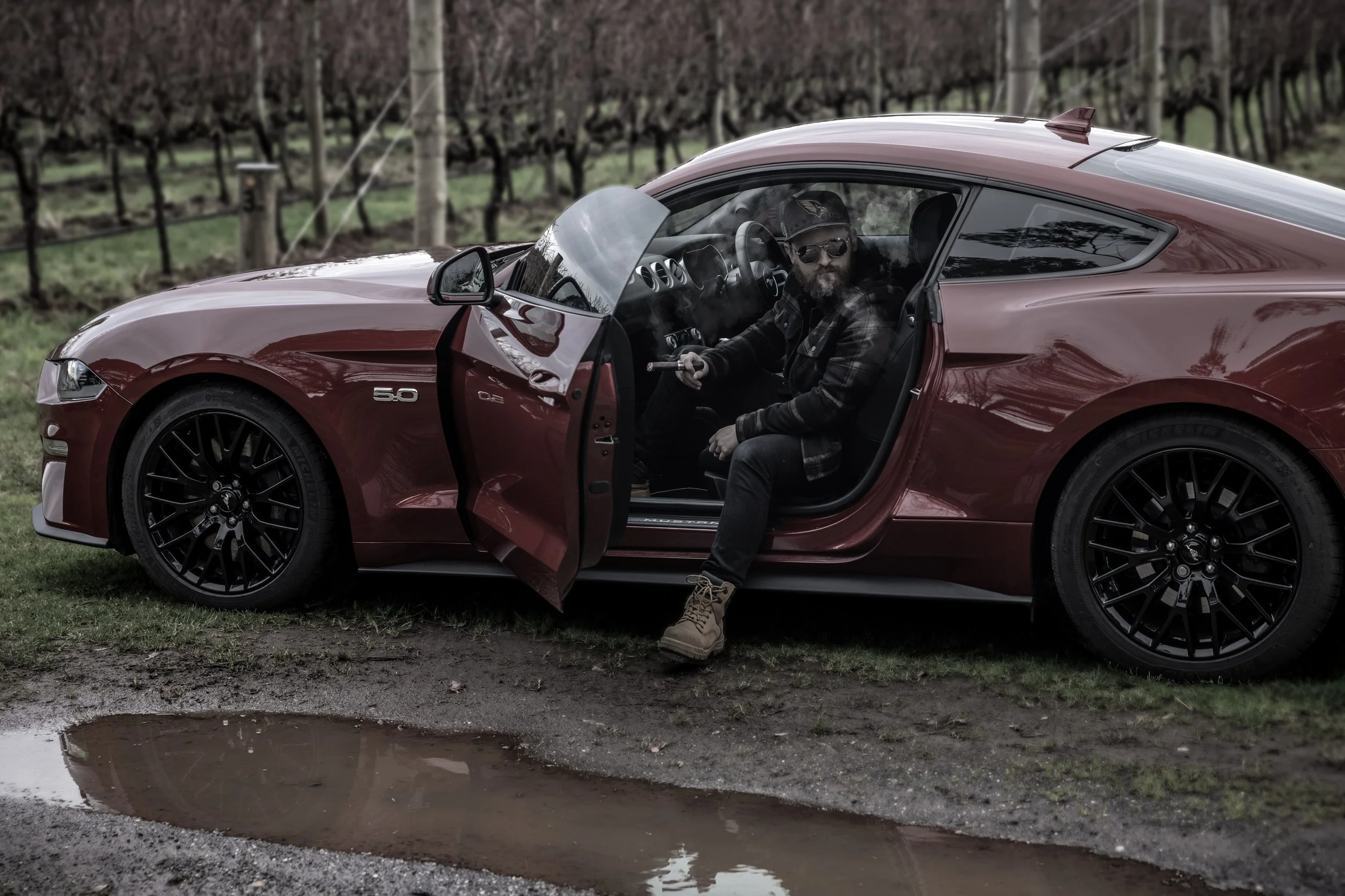 A man sitting in the driver's seat of a red Ford Mustang sports car with an open door, parked on a muddy area with a puddle in front. The background features a vineyard with rows of grapevines.