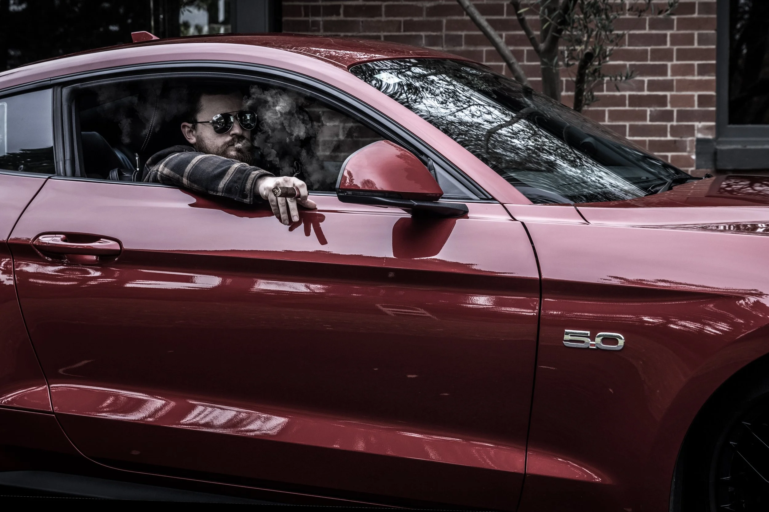 A man with sunglasses and a beard sitting in a red sports car, resting his arm on the window frame, with a brick building and trees reflected on the car's surface.