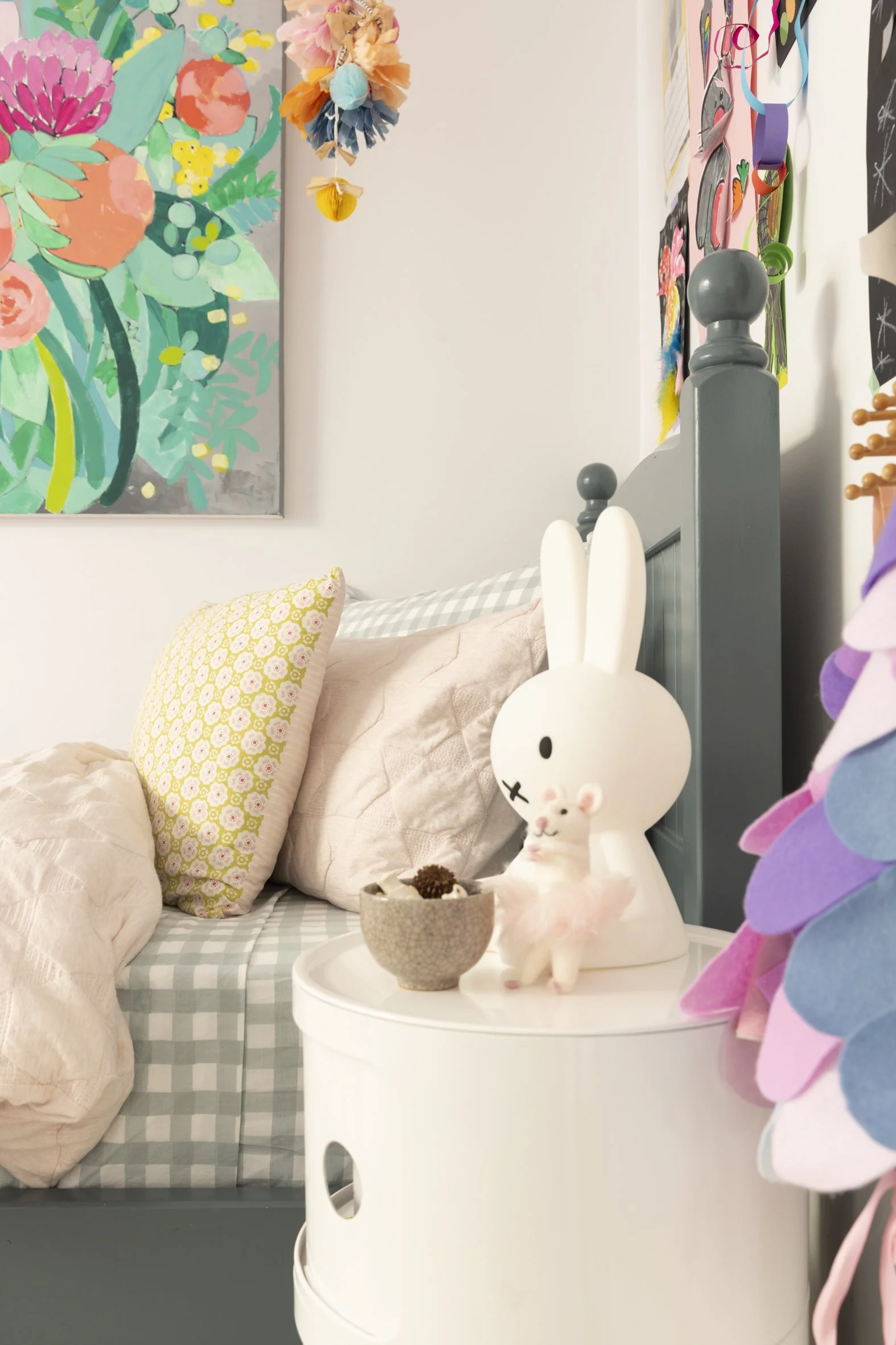 Children's bedroom with colorful floral painting, stuffed bunny toy on bedside table, checkered bedding, decorative pillows, and vibrant hanging decor.