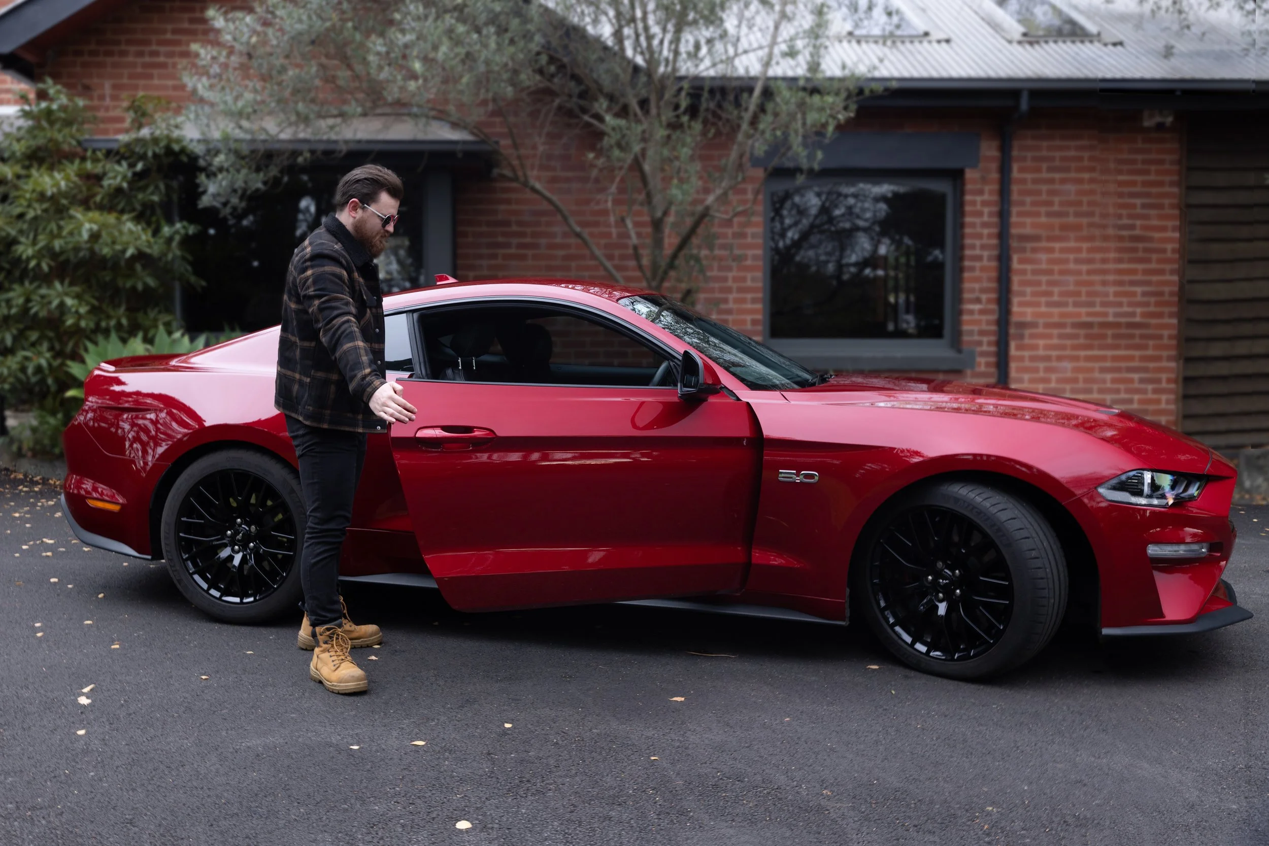 A man in sunglasses and a plaid jacket standing next to a red sports car with the driver's door open, in front of a brick house with trees.