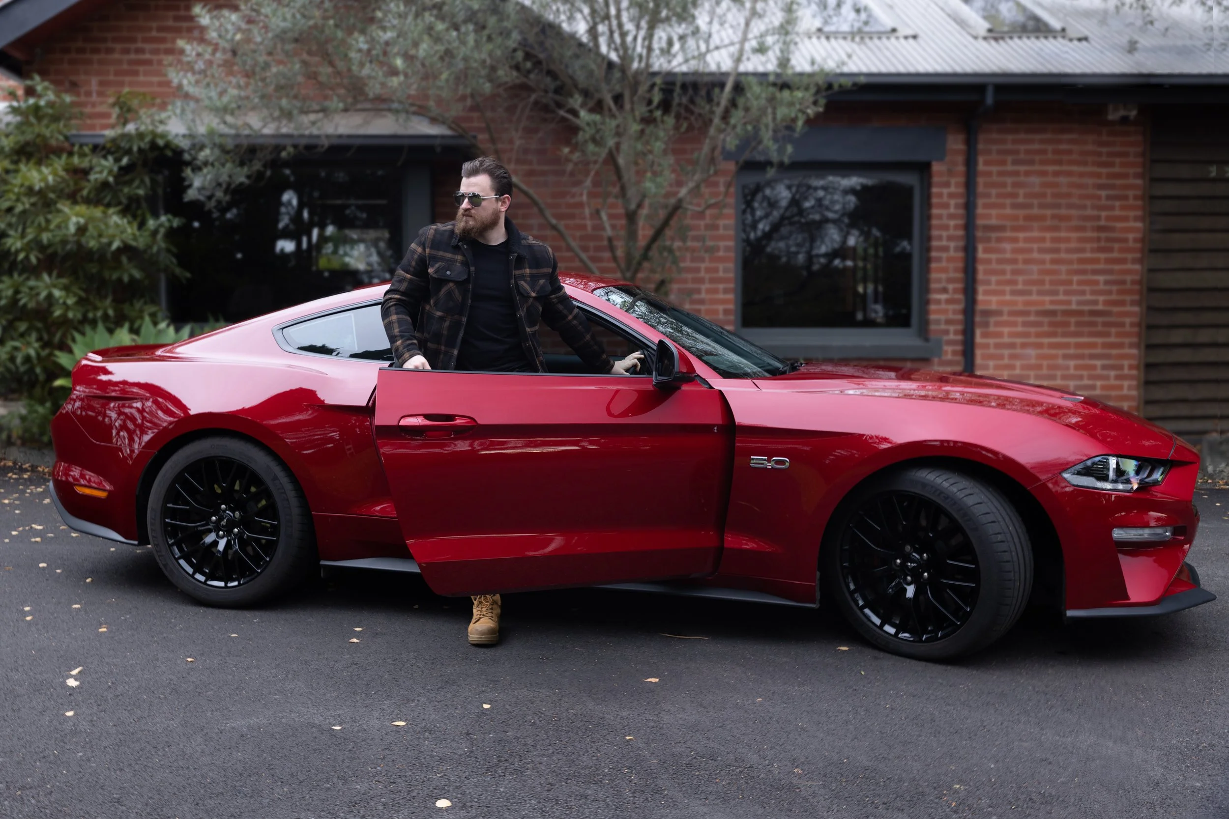 A man with sunglasses and a beard in a plaid jacket standing next to a red sports car with the driver’s side door open, in front of a brick house.