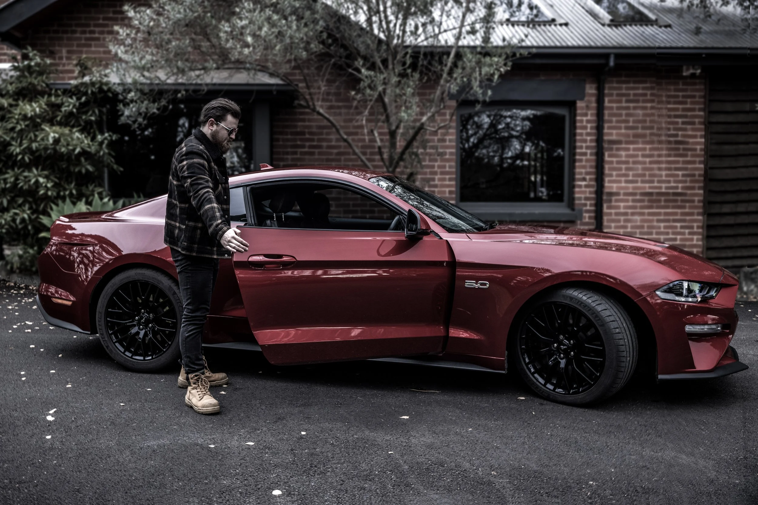 A man standing next to a red Ford Mustang with black wheels, parked outside a brick house, opening the driver's side door.