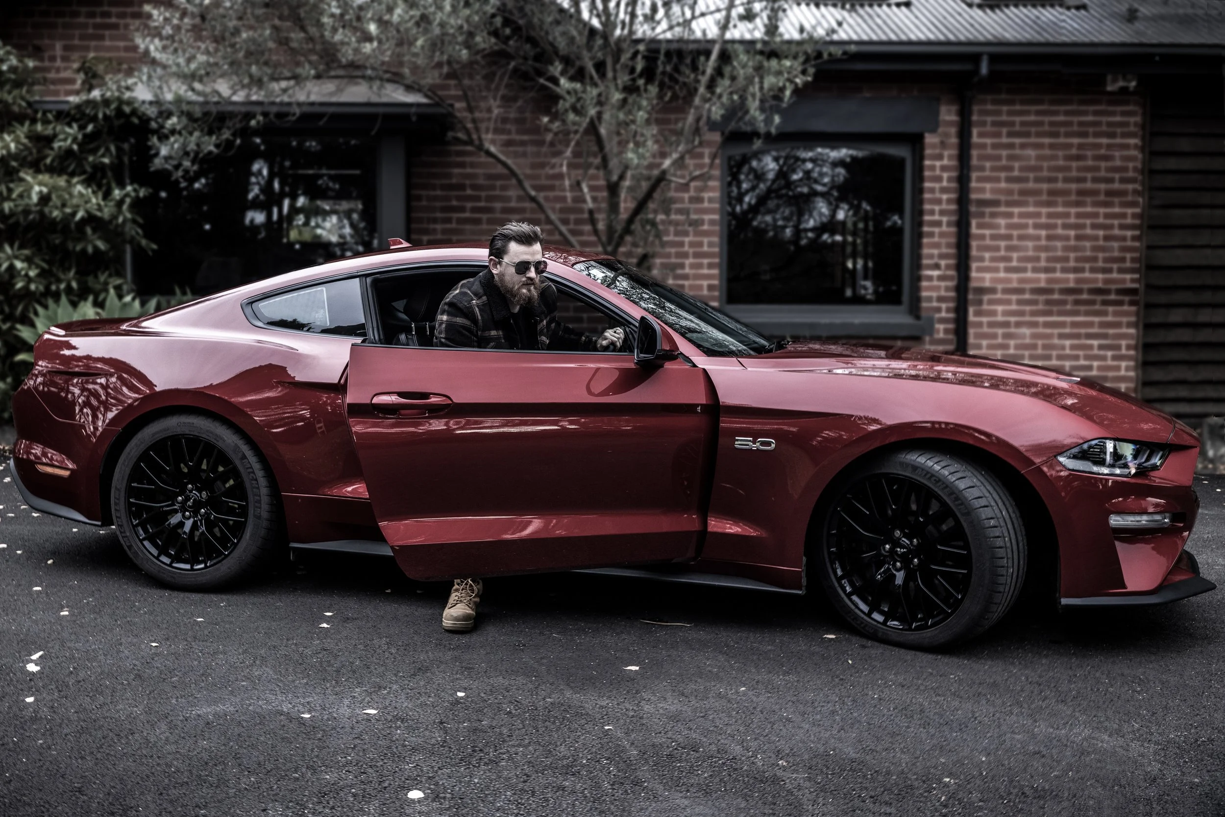 A man with sunglasses and a beard sits in the driver's seat of a red sports car with the door open, parked on a street in front of a brick building with trees and bushes nearby.