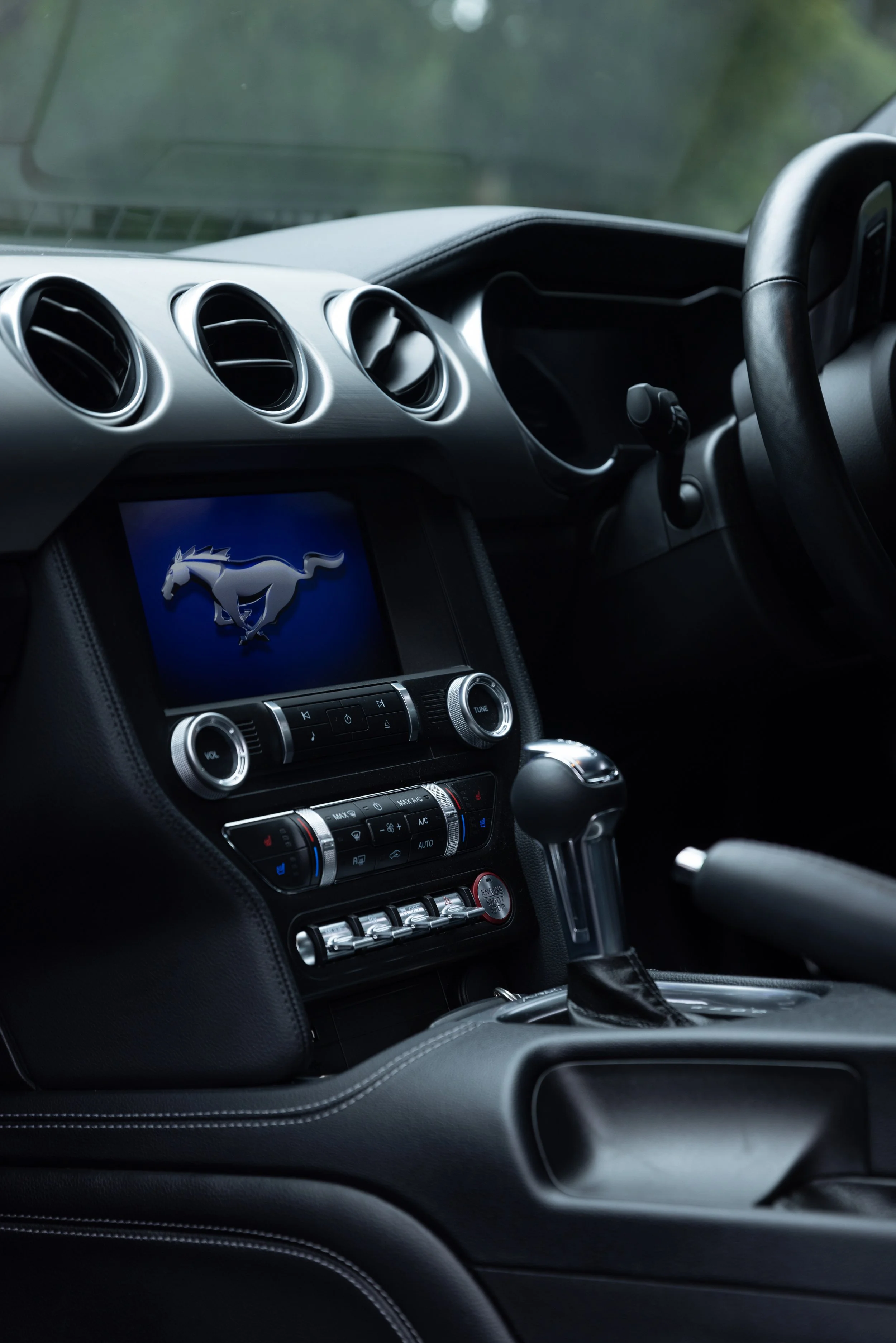 Interior view of a Ford Mustang car dashboard with a center console, leather-wrapped gear shifter, and a digital display showing the Mustang logo.