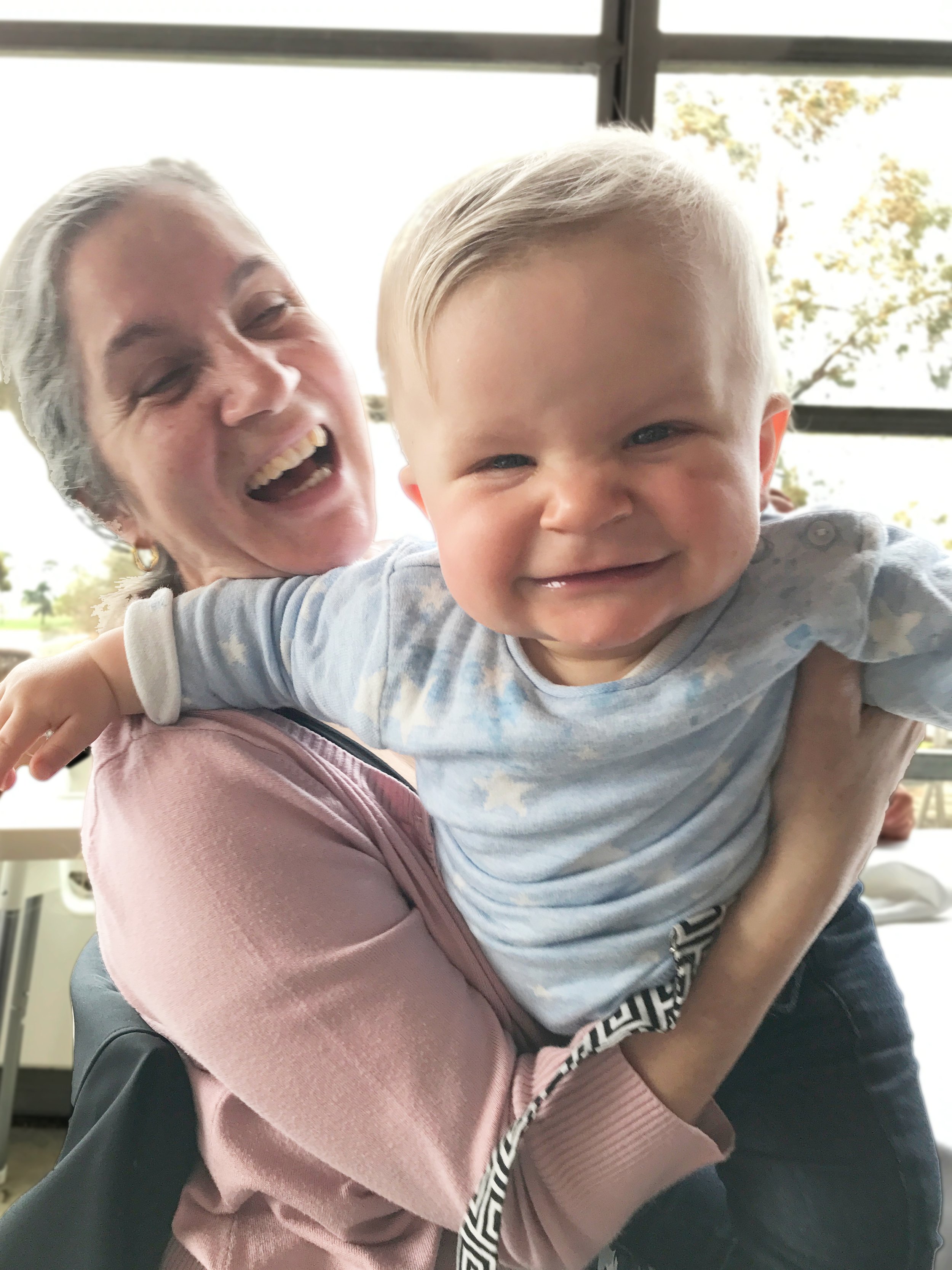 A Nonna holding her grandson, both appearing joyful indoors with large windows and trees outside in the background.