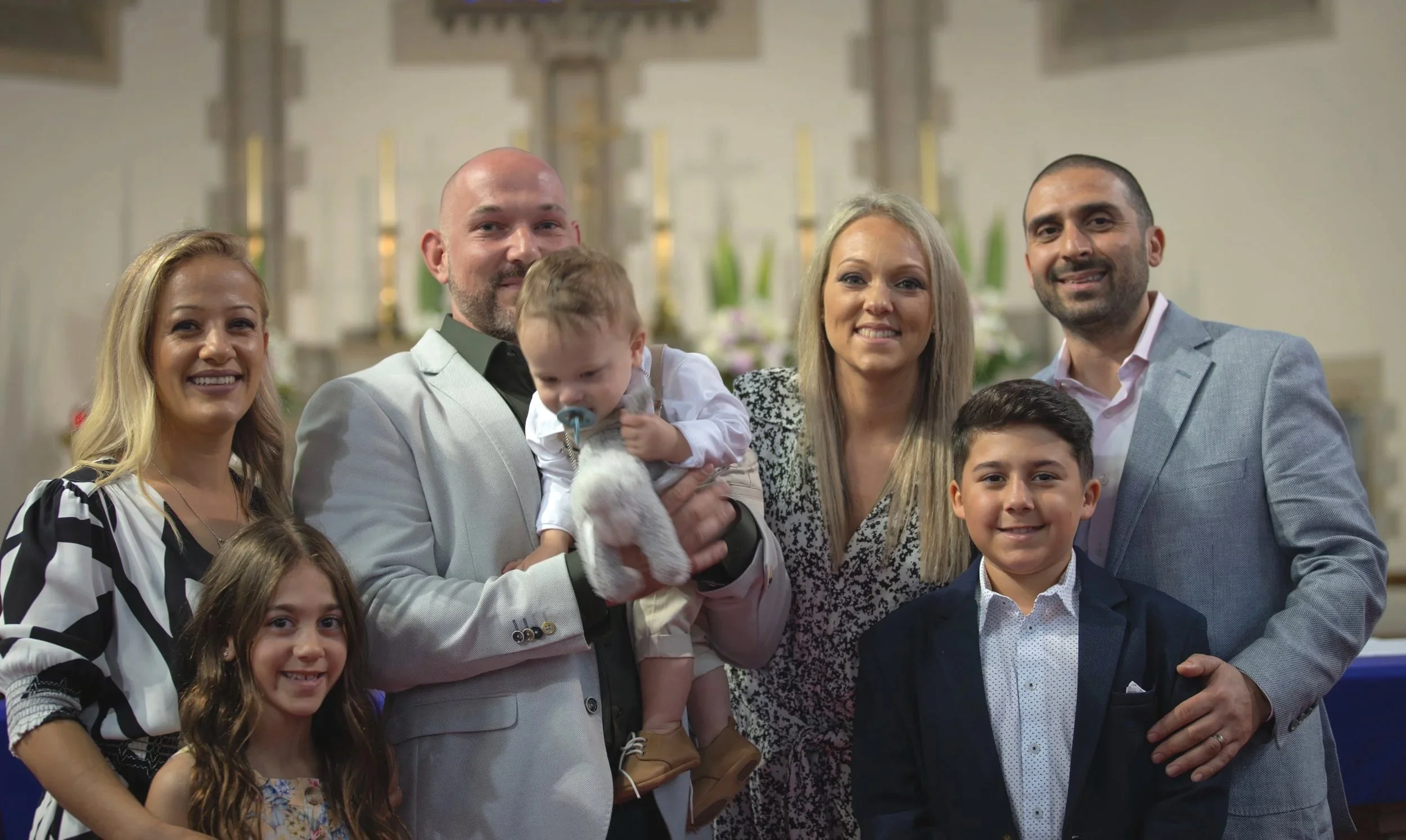 Group of eight people, including children and adults, smiling and posing together indoors, with a church altar in the background.