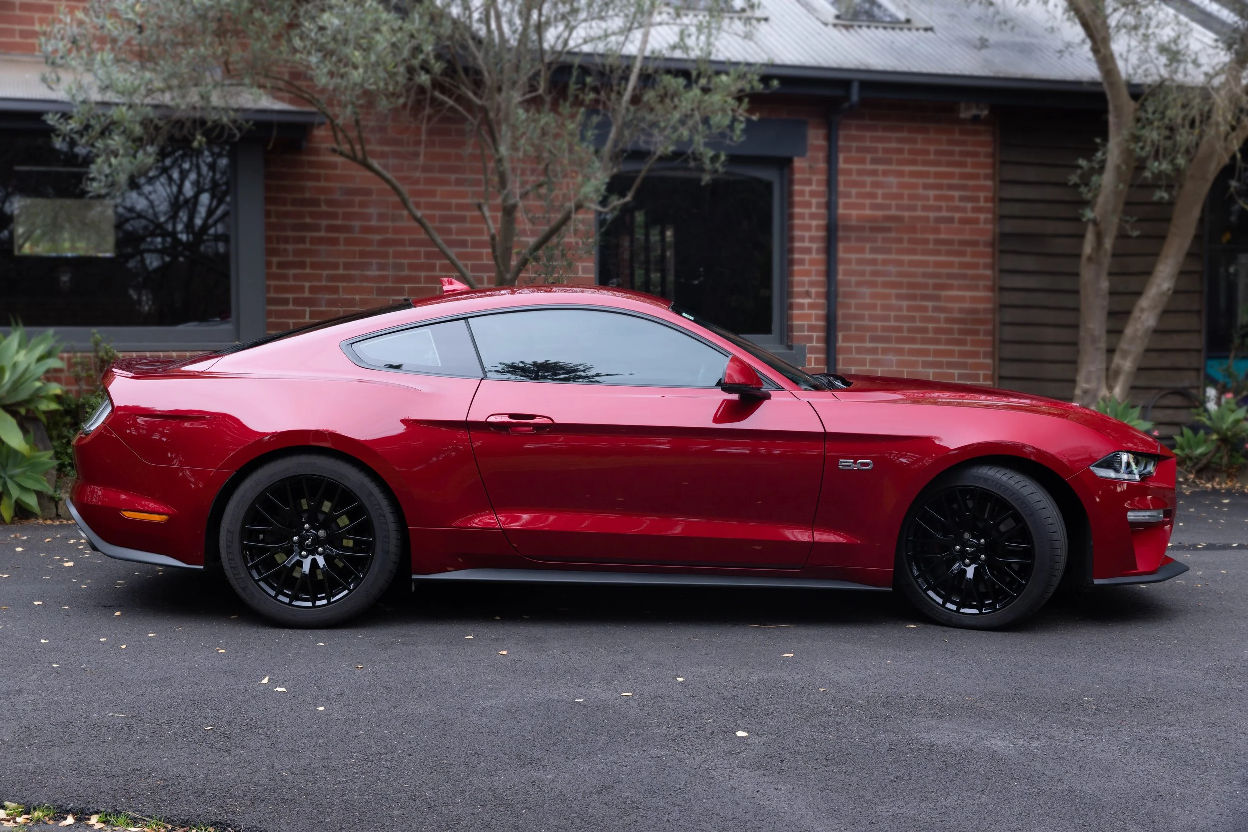 Red Ford Mustang sports car parked on a driveway in front of a house with brick and wood exterior and trees.
