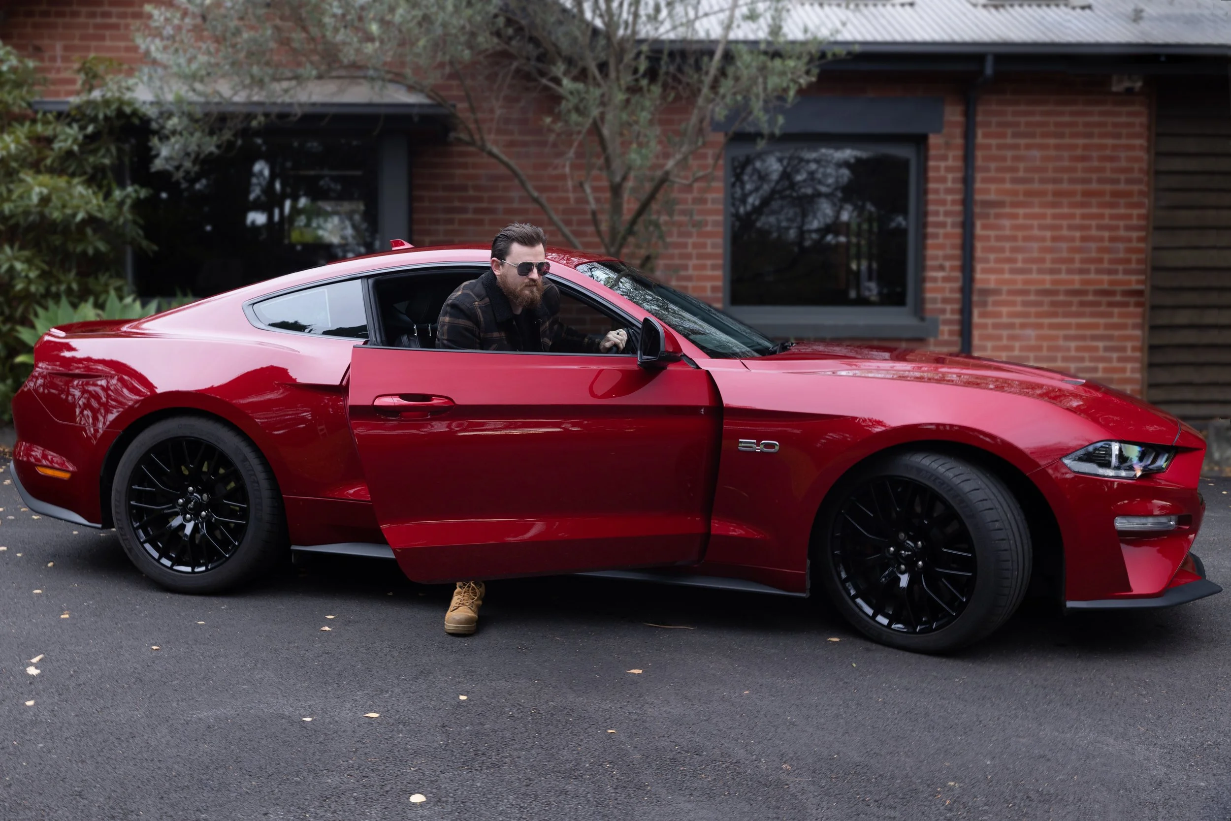 A man with a beard, sunglasses, and casual clothing sitting in a red sports car with the driver's door open, parked on a residential street with brick houses and trees in the background.