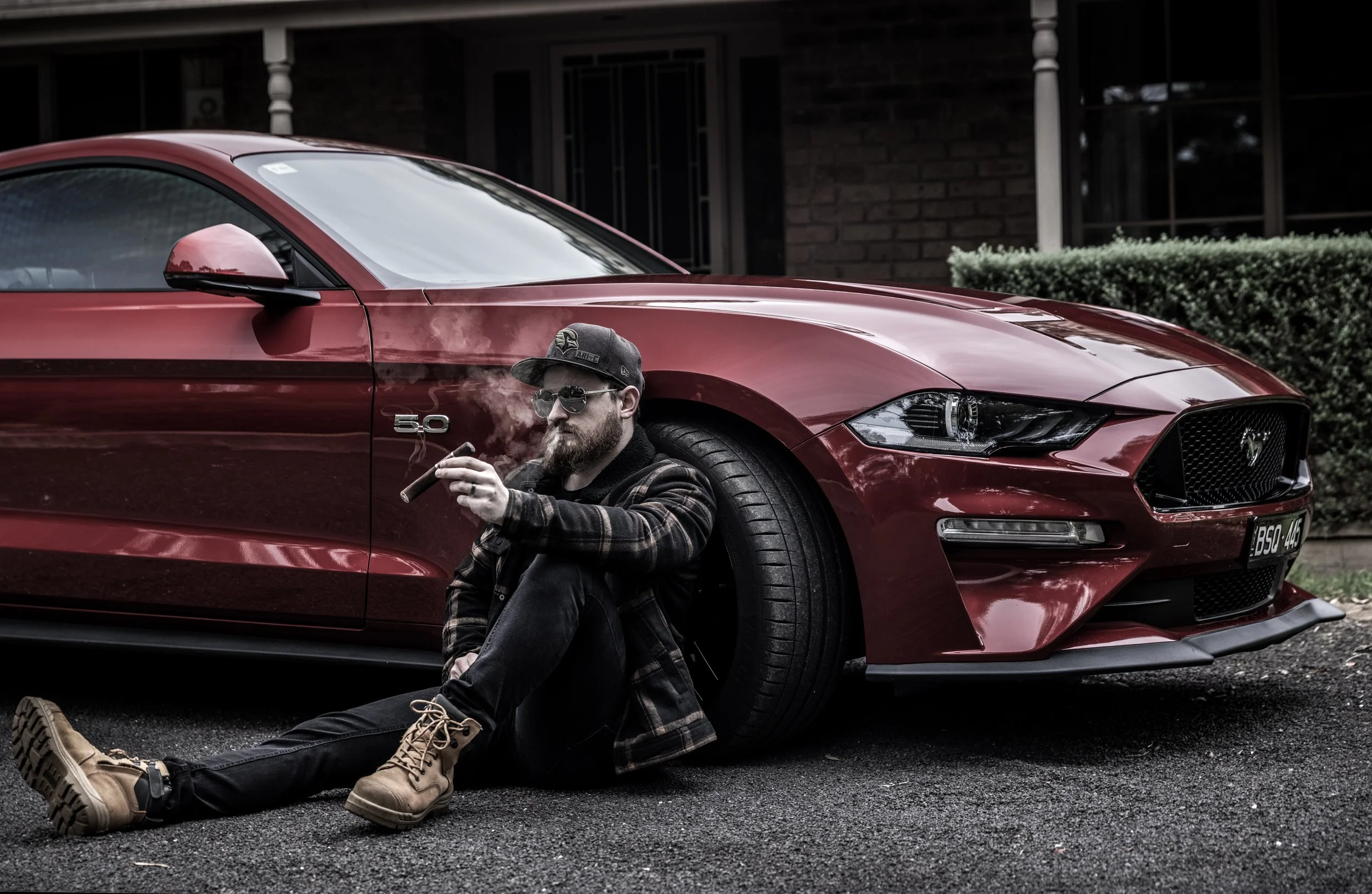 A man with a beard and sunglasses sitting on the ground next to a red Ford Mustang, smoking a cigar. He is wearing a cap, a plaid jacket, and tan boots.