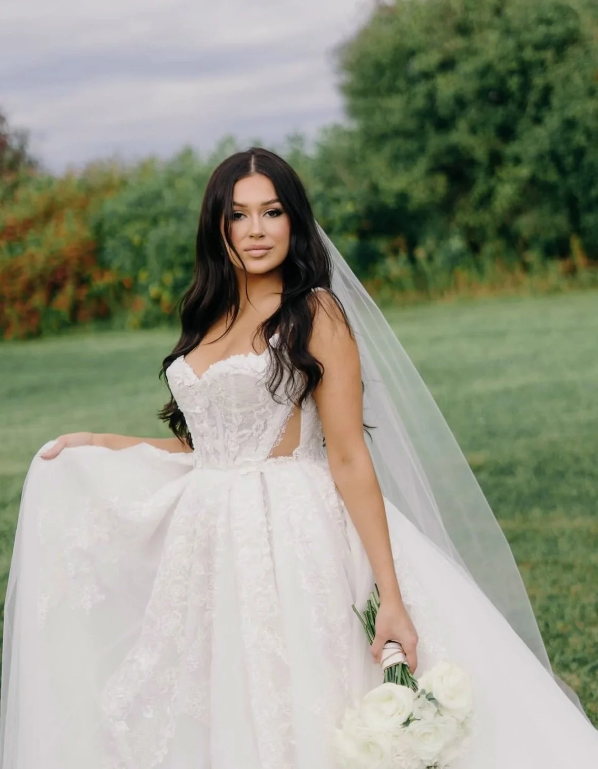 A woman in a white wedding dress holding a bouquet of white roses outdoors with green trees in the background.