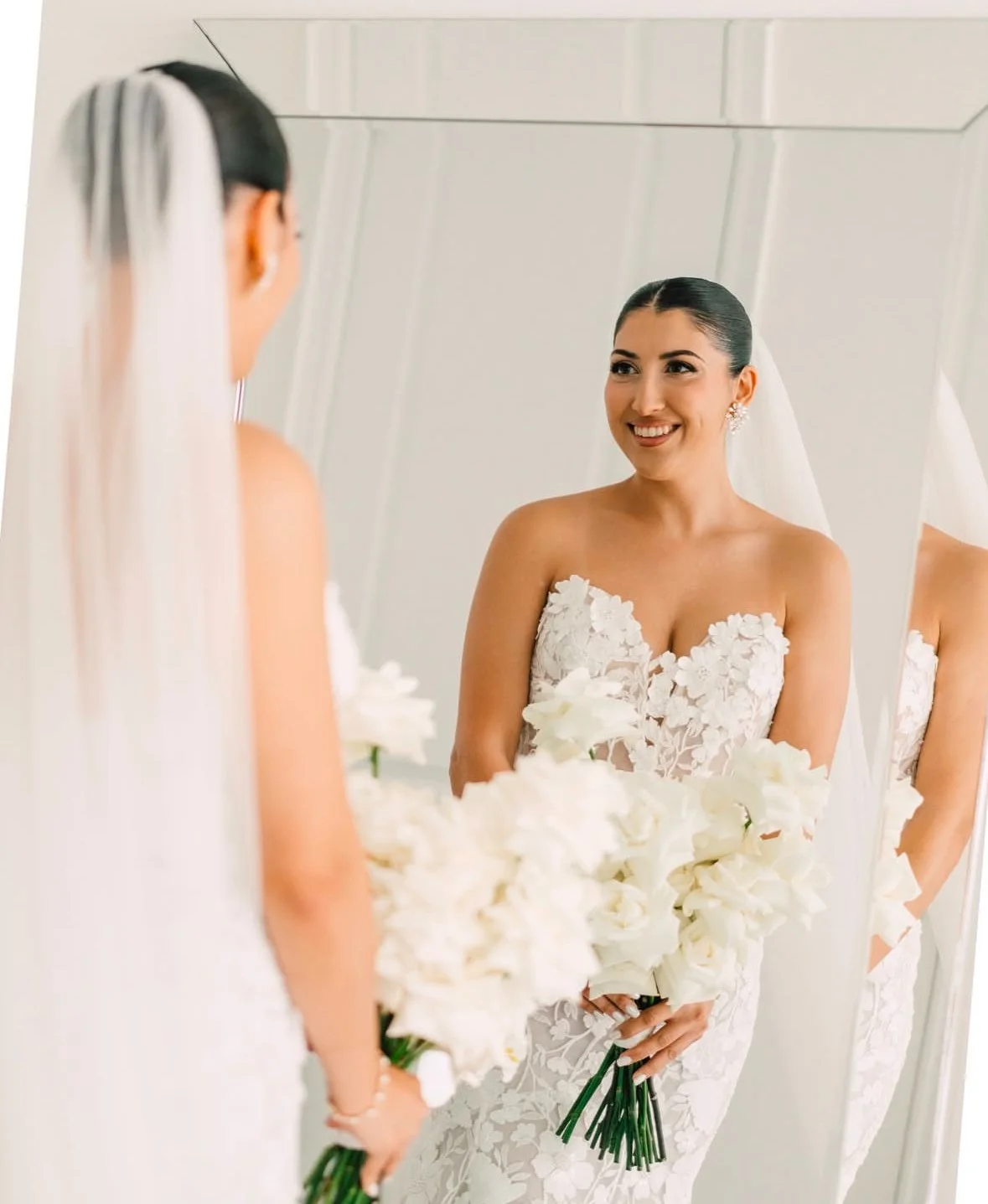 A bride in a white strapless wedding dress with floral details, holding a bouquet of white flowers, smiling at her reflection in a mirror.