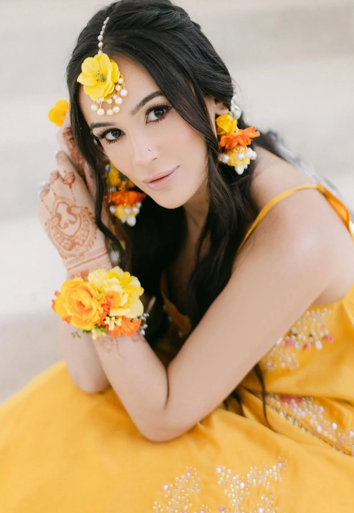 A woman dressed in a yellow traditional outfit with floral jewelry and henna on her hand, posing with her arms crossed in front of her face.