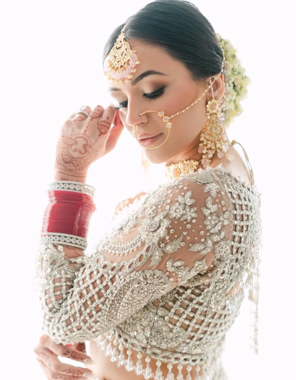 A bride in traditional Indian attire with intricate jewelry and henna on her hand, posing with her eyes closed and a gentle expression.