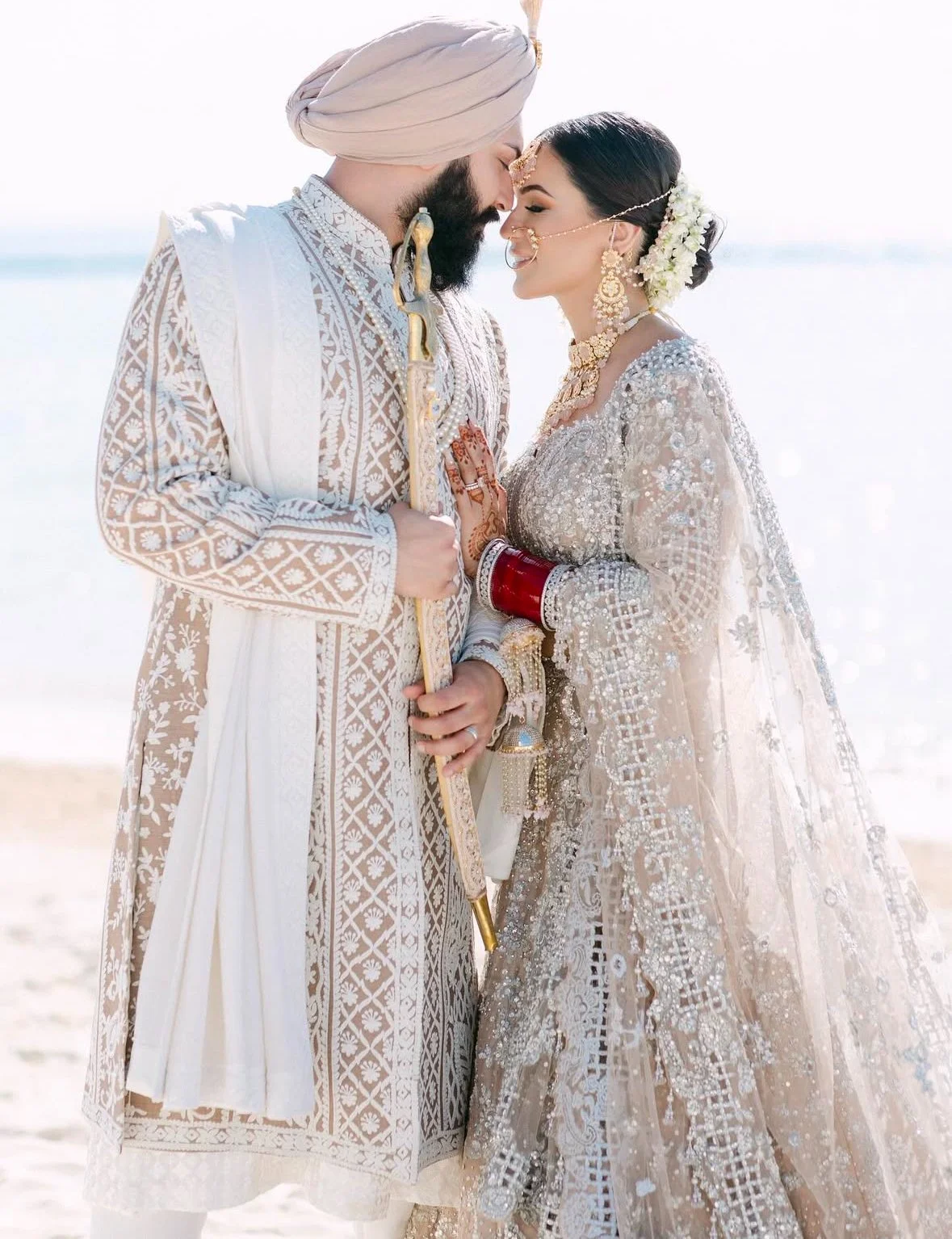 A bride and groom in traditional Indian wedding attire at the beach, gently touching foreheads with eyes closed.
